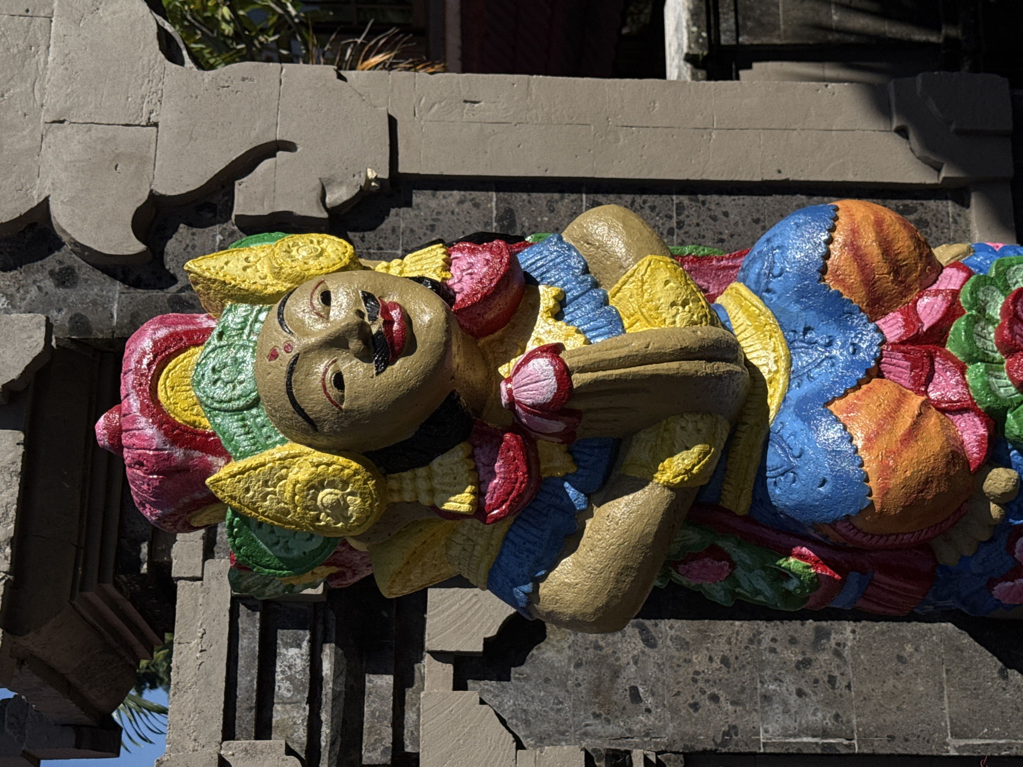 A brightly painted, colorful statue stands in front of a stone structure, likely in the city of Ubud District, Indonesia. The statue depicts a human-like figure with a golden-brown face, red lips, and a black mustache. The figure wears a blue garment over a patterned orange lower body, adorned with red and pink floral accents. Elaborate headdresses and shoulder decorations in yellow, green, and red surround the figure's head and upper body. The statue's hands are clasped together in front of its chest, holding a pink lotus-like flower. The stone structure behind the statue is made of dark, rough-hewn blocks, with a lighter, more decorative stone element visible to the right. Sunlight casts shadows on both the statue and the stone architecture, indicating an outdoor setting. A hint of blue sky is visible above the structure on the left.