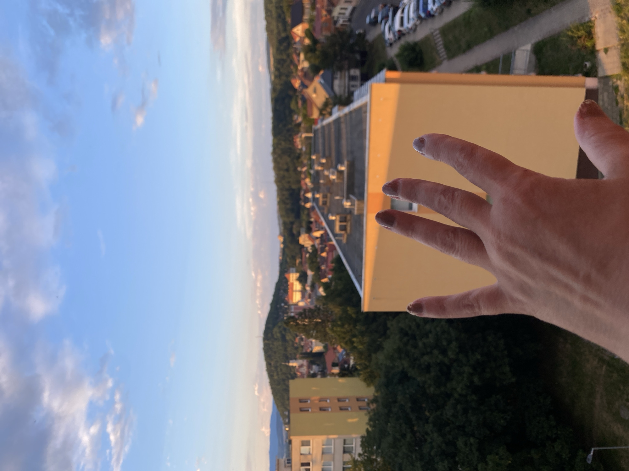 An overhead, eye-level shot captures a woman's hand, fingers outstretched, as she poses in front of a cityscape at sunset. The hand is positioned in the foreground, obscuring the lower portion of the image. The cityscape reveals buildings, trees, and a distant hill. The lighting suggests it's either dawn or dusk. The overall impression is one of a scenic overlook in Bílina, Czechia.