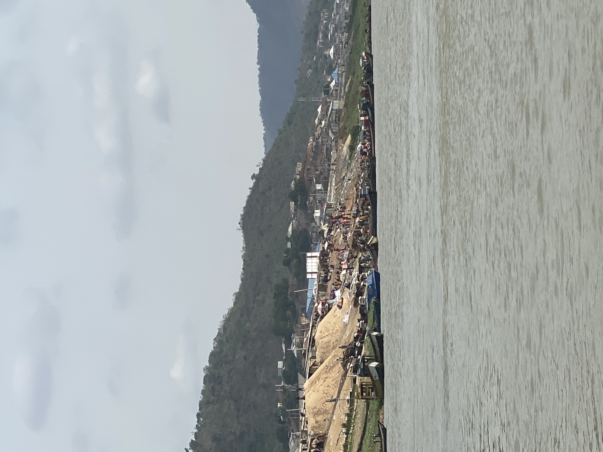 The image shows a waterfront scene in Lokoja, Nigeria. A wide river occupies the right side of the frame. The left side features a steep hillside densely populated with buildings and structures, suggesting a settlement. At the base of the hill, alongside the river, is a sandy area possibly used for construction or industry, with equipment and structures visible. People are present on the sandy area and amidst the buildings, though their activities are not clearly discernible. The sky is overcast, and a mountain range is visible in the background, further inland.