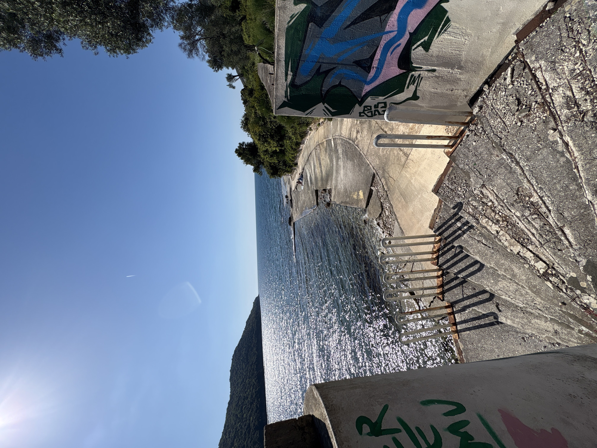 Captured from a high vantage point in Bratica - Braticë, Montenegro, the image presents a striking landscape where architecture meets the sea. The foreground consists of weathered concrete structures bearing graffiti. Stairs descend towards the shimmering water. The sea stretches out with sunlight creating a glittering surface. A verdant landmass flanks the water's edge. The sky is a clear blue with a faint vapor trail, contrasting with the rugged concrete.