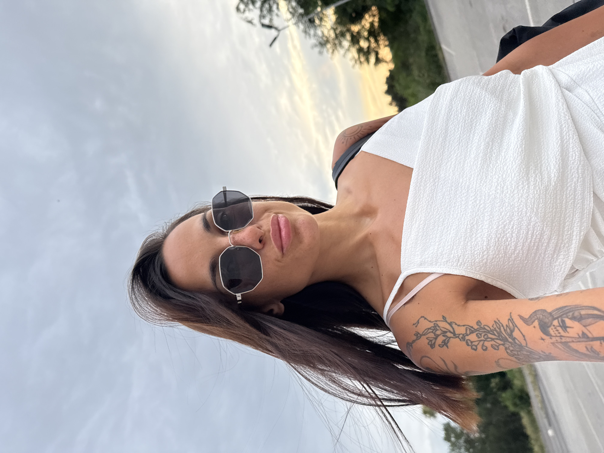 An outdoor, eye-level, medium-full shot captures a woman posing for a selfie on a cloudy day. The woman, with dark brown hair that is blowing in the wind, wears large, dark, octagonal sunglasses and a textured white top. She has tattoos on her left arm and shoulder. The background consists of a gray sky with some lighter clouds, trees, and a street lamp. The scene appears to be an outdoor parking lot, possibly in the city of Pleven, Bulgaria, given the presence of trees and a street lamp common in urban environments.