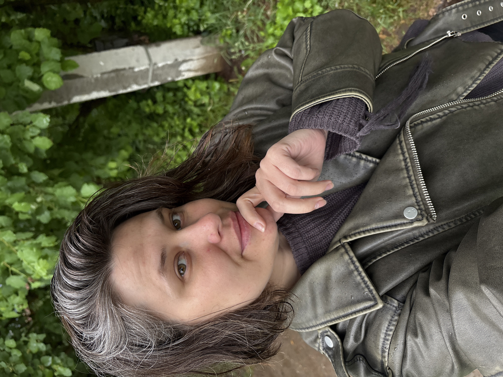 A woman with shoulder-length, dark brown hair with streaks of gray posing for a selfie. She is wearing a gray sweater and a distressed, olive green leather jacket with silver zippers and buttons. Her hand is lightly touching her chin. The background is blurry, showing green foliage and a concrete structure, suggesting a park or natural area. The photo was likely taken outdoors in natural light. The location is Chaikovsky, Russia, as indicated by the prompt.