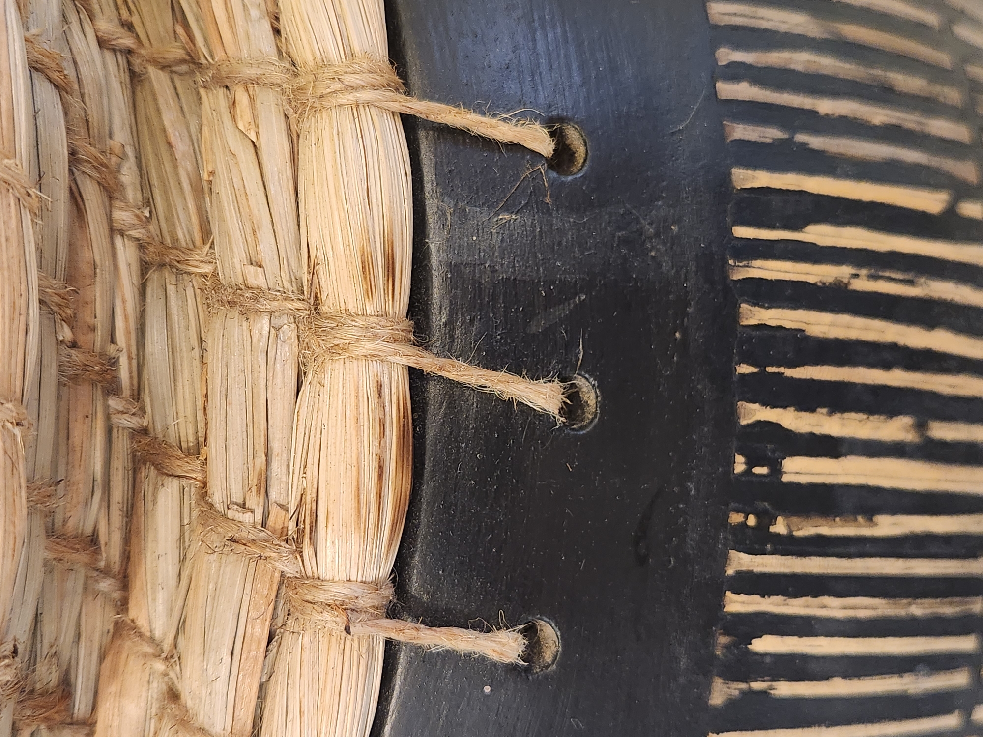 This is a close-up, eye-level, horizontal shot of a textured object, likely a drum or shield, due to the presence of woven straw and what appears to be a dark, possibly wooden or leather surface. The straw is bundled together and secured with rough twine in several places. The twine is tied around the bundles and then extends to the dark surface, where it passes through drilled holes. Three such ties are visible, with the twine ends frayed. The dark surface shows two circular indentations where the twine passes through. To the right of the dark surface, there is a section with alternating bands of dark material and lighter material that appears to be the same straw or a similar natural fiber. These bands are oriented horizontally in the image. The lighting is even, suggesting it was taken indoors or in overcast conditions, with no strong shadows. The overall impression is one of rustic craftsmanship and natural materials. No people, text, or specific location cues are visible.
