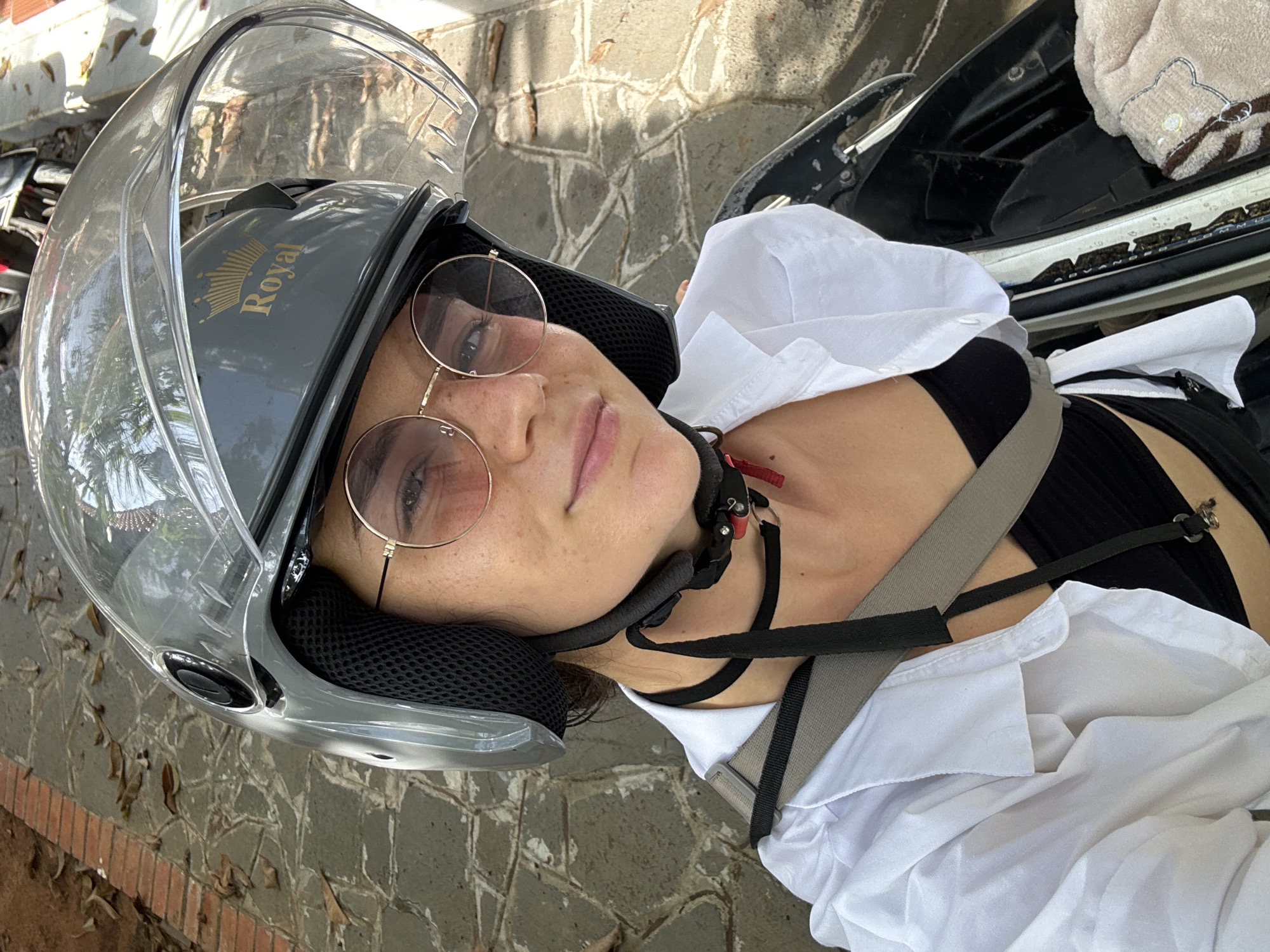 A woman wearing a gray motorcycle helmet with the word "Royal" on it, along with round, gold-rimmed glasses, is lying on a gray brick surface in an outdoor setting. The surface has fallen leaves scattered around. She is wearing a white button-down shirt over a black top and a black choker. A gray strap is positioned across her body. The background shows sections of the motorcycle she is presumably riding. Based on the general appearance and style, the location may be Mũi Né, Vietnam.