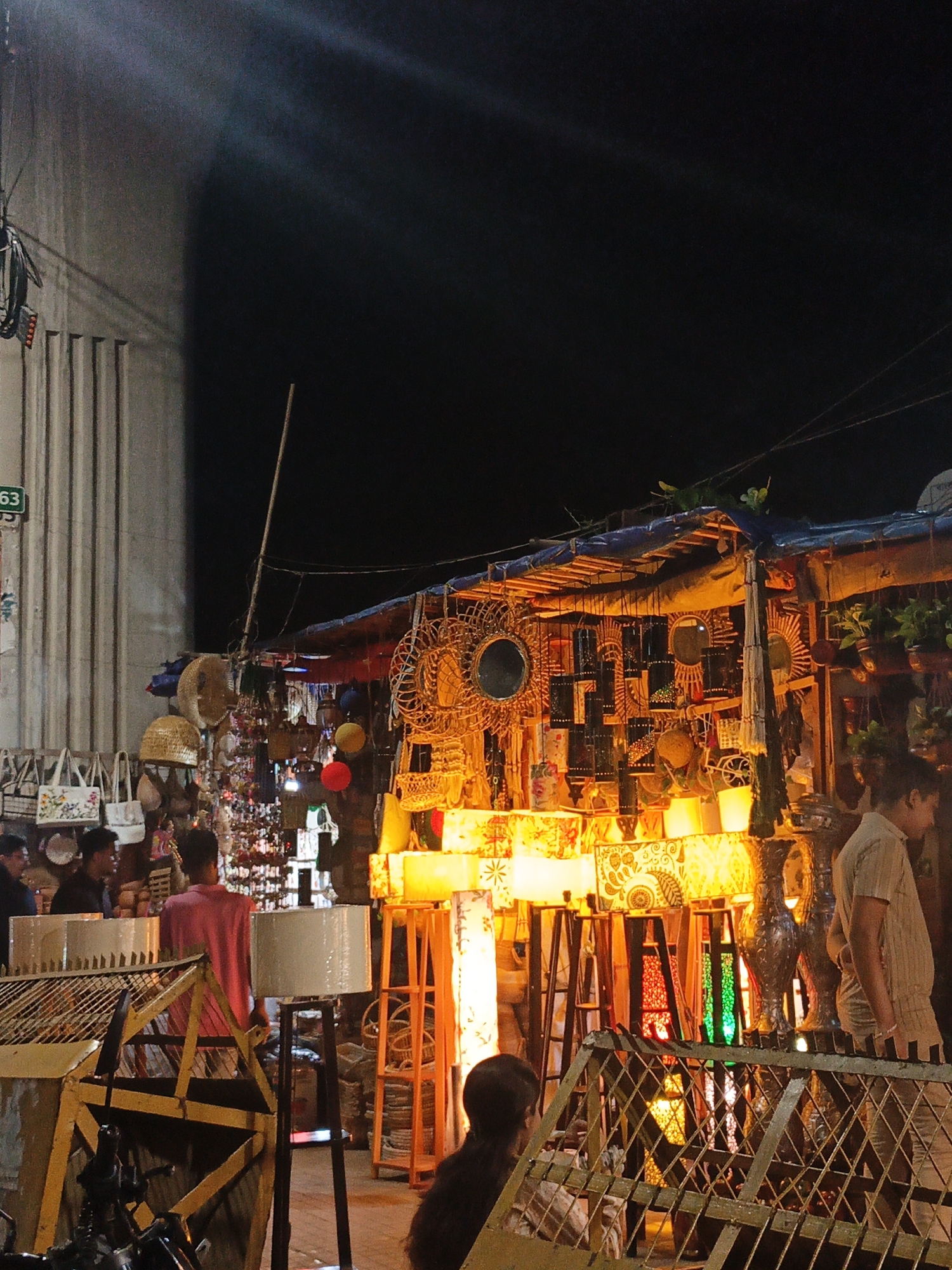 The image depicts a night scene at an outdoor market stall in an unknown city. The stall is illuminated by various lamps and colorful lights, showcasing an array of handcrafted items, including woven baskets, decorative mirrors, and lampshades with intricate patterns. Several individuals are visible, some browsing the merchandise and others attending to the stall. The background features a large, ribbed concrete structure and a dark night sky. The overall impression is one of a vibrant, bustling marketplace at night.