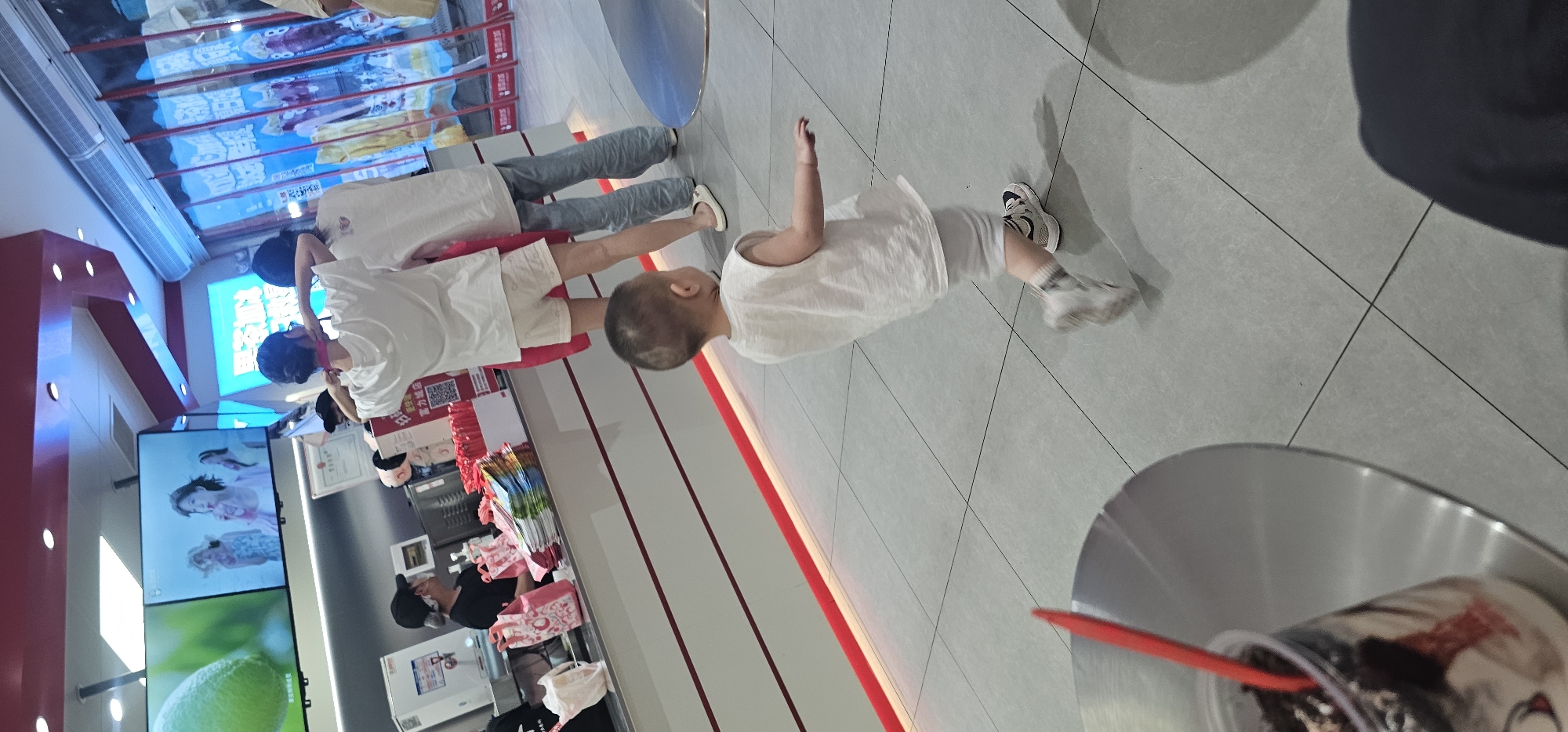 In a brightly lit indoor commercial establishment, possibly a fast-food restaurant or shop, a young child wearing a white sleeveless shirt and shorts runs across a tiled floor. The child is in motion, with their left arm extended and right arm bent. They are wearing white sneakers with black and grey accents. In the foreground, to the left, a portion of a metal table is visible with a clear plastic cup containing a dark-colored beverage and a red straw. In the background, a counter area displays various items, including pink shopping bags, colorful packaged products, and what appear to be promotional materials on screens. A few individuals, including a person wearing a black cap and apron, are visible behind the counter, and other customers are standing in a line. The environment suggests a retail or service setting within the city of Chongqing High-tech Zone, China, given the context.