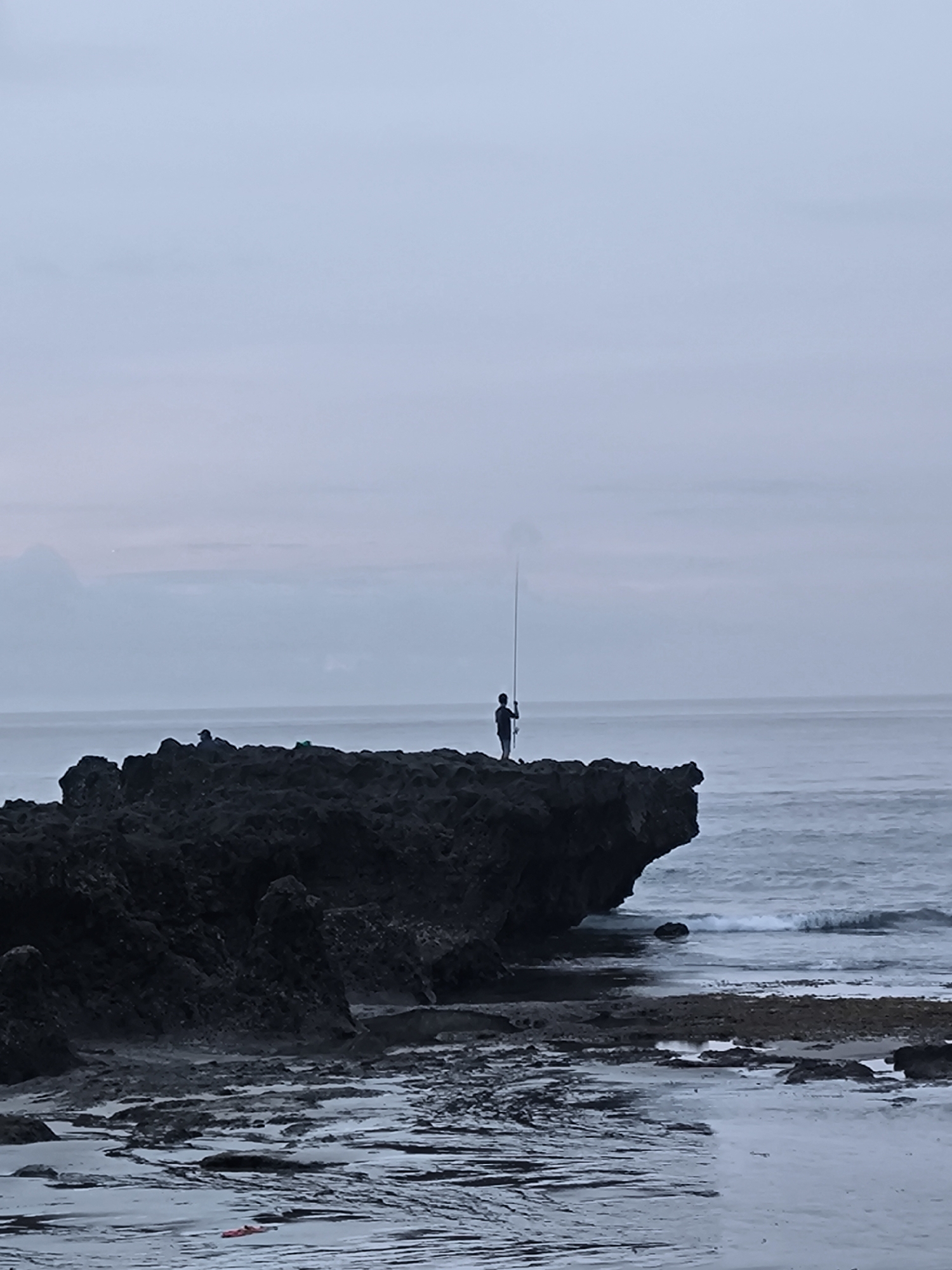 A figure stands on a dark, rocky outcrop extending into the ocean, holding a fishing rod. The ocean is visible in the background, with a calm surface and some wave action near the rocks. The sky is overcast, suggesting a cloudy or dusk-like environment. The foreground includes tidal pools or wet sand, reflecting the sky.