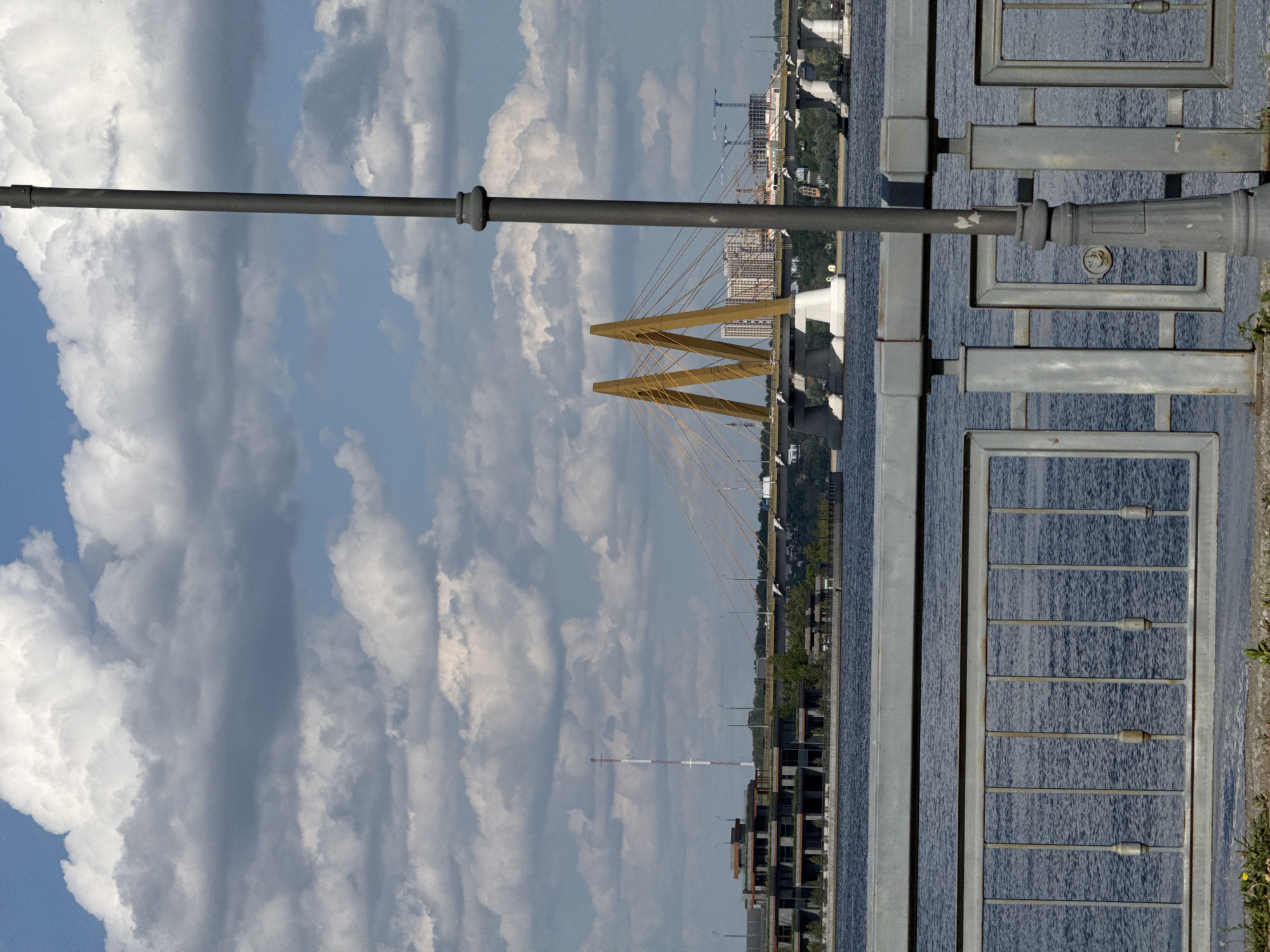 The image shows a view of Kazan, Russia, featuring the Millennium Bridge. The bridge, a prominent landmark, is a cable-stayed bridge with a distinctive M-shaped yellow pylon. The bridge spans the Kazanka River, visible as a dark expanse running through the scene. In the background, a cityscape emerges, with buildings, cranes, and construction sites indicating urban development. An industrial chimney with red and white stripes is also visible. The foreground is defined by a gray metal railing and a lamppost, suggesting the viewpoint is from a sidewalk or pedestrian area. The sky above is filled with puffy, partly cloudy formations, providing a bright, daytime setting. The image is oriented sideways, so elements on the right side of the photo may appear upside down.