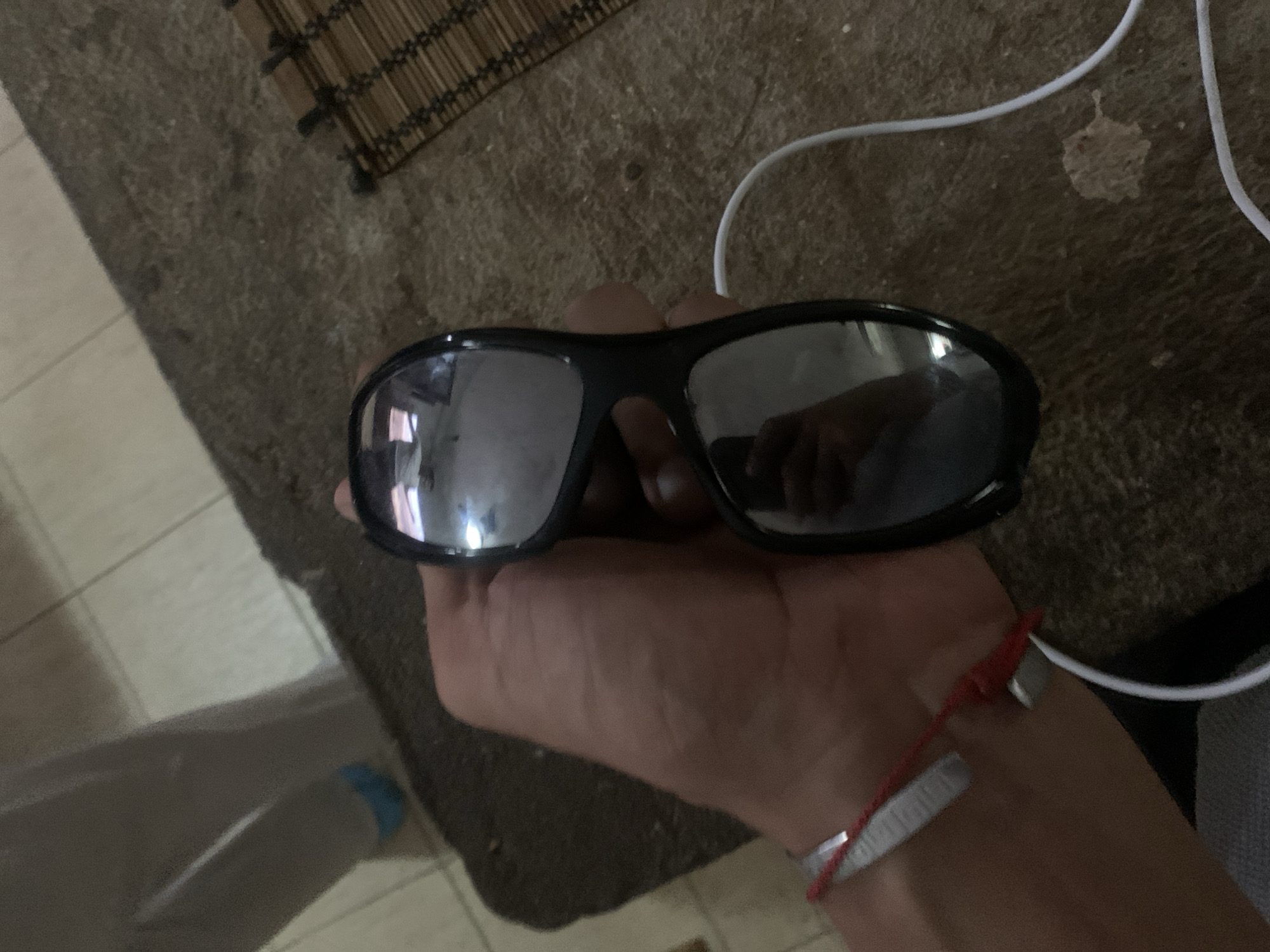 The image shows a hand holding a pair of black framed sunglasses with reflective lenses. The background consists of a textured rug, a rolled-up bamboo mat, and a white cable. The person is wearing a red string bracelet with metallic beads.
There is no indication of the city Santa Ana, Venezuela, or its environment in this image.