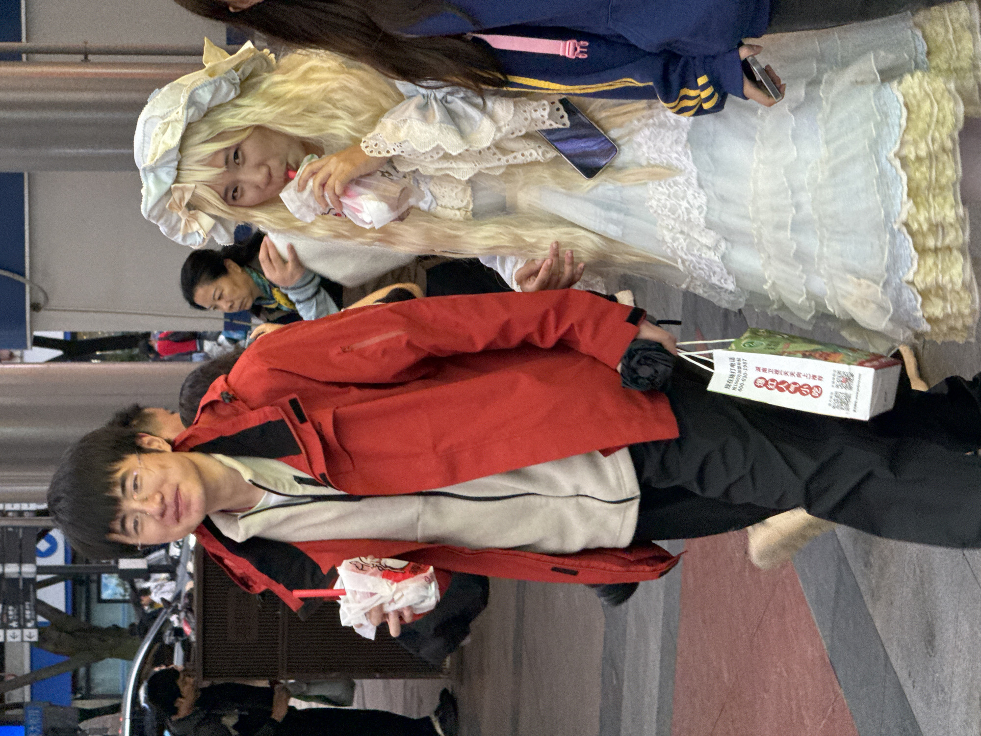 In Jinjiang District, China, a group of people are visible. A person with black hair, glasses, and a red jacket holds a white object with a red stick. Adjacent to this person is another individual wearing a frilly white dress and a white bonnet, also holding an object. There is another person nearby. A printed box is held by the person in the red jacket with Chinese text visible. The environment appears to be a street scene with urban infrastructure such as poles and street signs. The ground is paved with gray and red tiles. A phone is in the hands of the person in the frilly white dress.