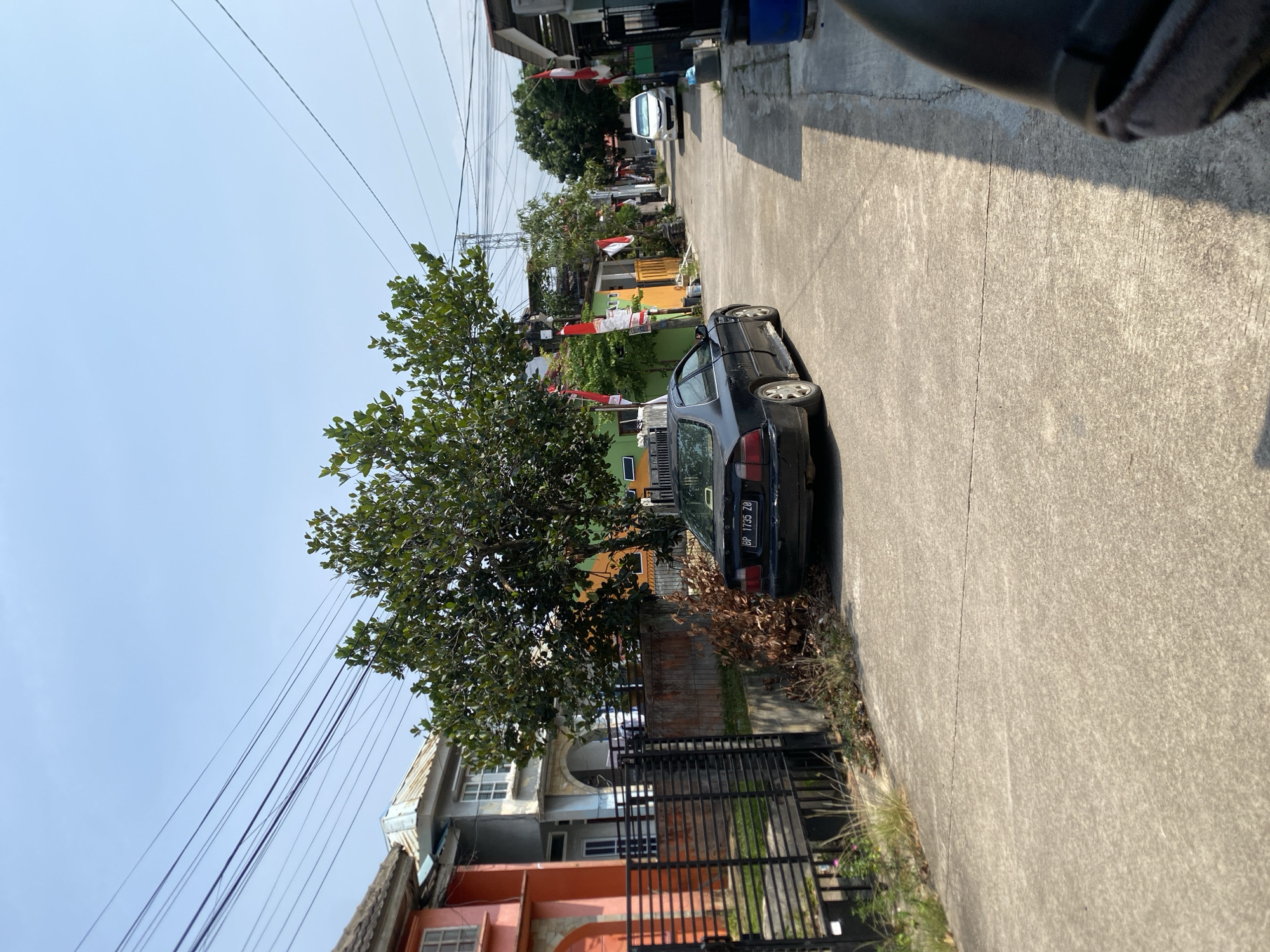 A daytime, eye-level shot shows a residential street in Batam, Indonesia. The street is paved with concrete and lined with houses, some of which are painted in bright colors like orange and yellow. Several trees with lush green foliage are scattered throughout the scene, providing shade. Various power lines crisscross overhead. In the foreground, a black sedan is parked on the side of the road, displaying a license plate that reads "BP 1735 ZO". A white car is visible further down the street. Indonesian flags are displayed on poles in front of some houses, suggesting a celebration or national holiday. The sky is clear and blue.