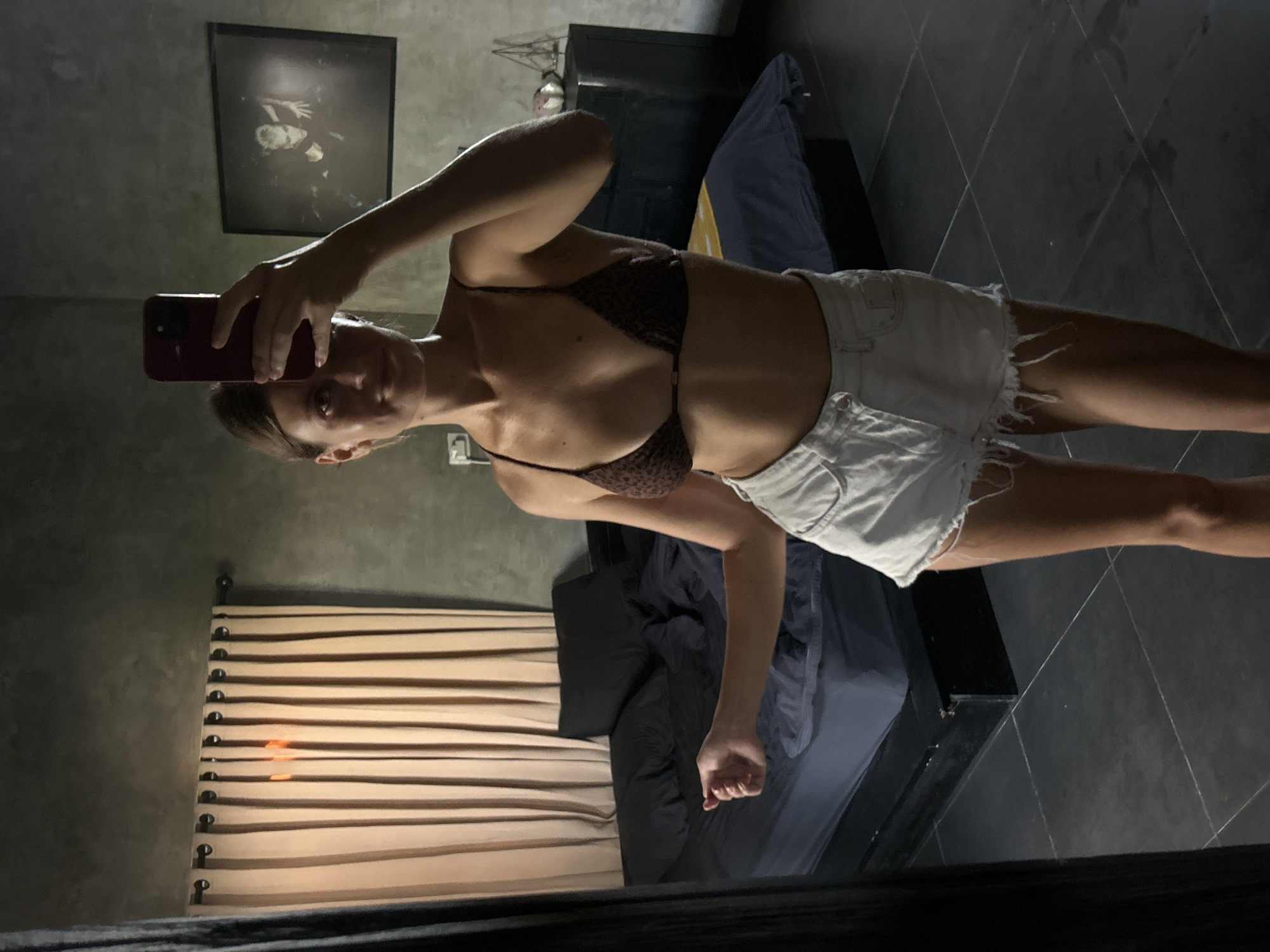A woman with brown hair is taking a selfie in a room. She is wearing a brown patterned bikini top and white denim shorts. The room has a dark aesthetic with grey walls, a dark bed frame, and black tiled floors. A framed picture hangs on the wall. Wooden blinds are visible on the left side of the image. The location appears to be indoors, but the specific details do not confirm it is Mengwi, Indonesia. The woman's action is taking a selfie with her phone.