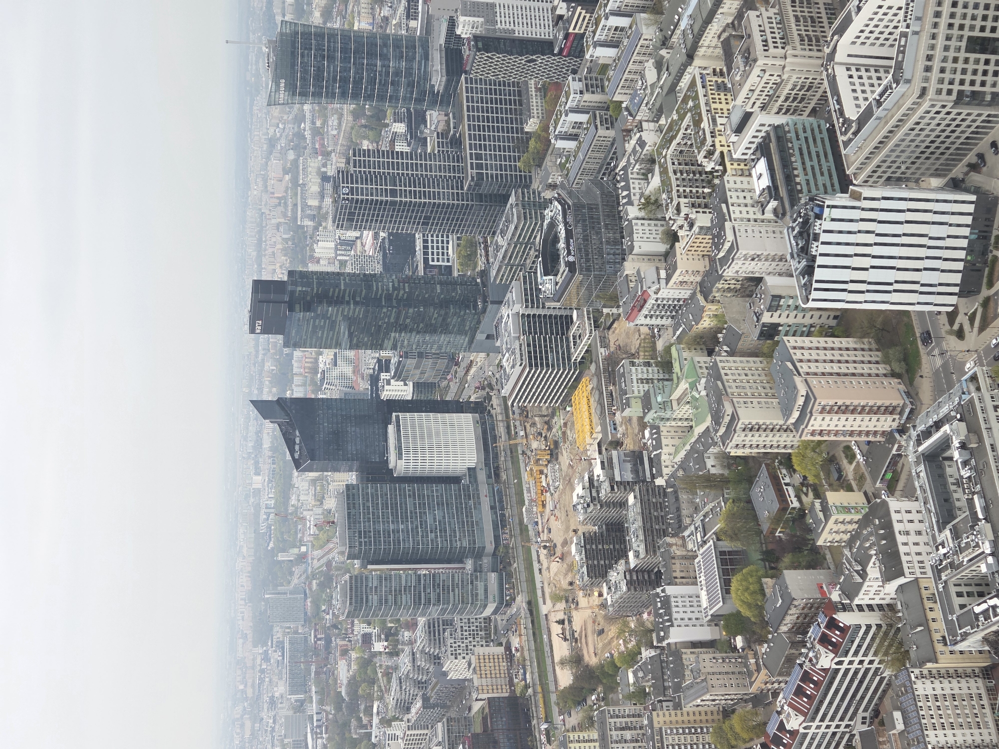 The image shows an aerial view of Warsaw, Poland, with a focus on the city's downtown area. Several tall skyscrapers dominate the skyline, some bearing logos like "Samsung" and "Hertz". Construction sites with yellow cranes and various construction vehicles are visible along a central corridor, indicating ongoing development. The city is a mix of modern high-rises and older, low-rise buildings. The overall environment is urban, densely populated, and slightly hazy. No people or specific actions are readily apparent.