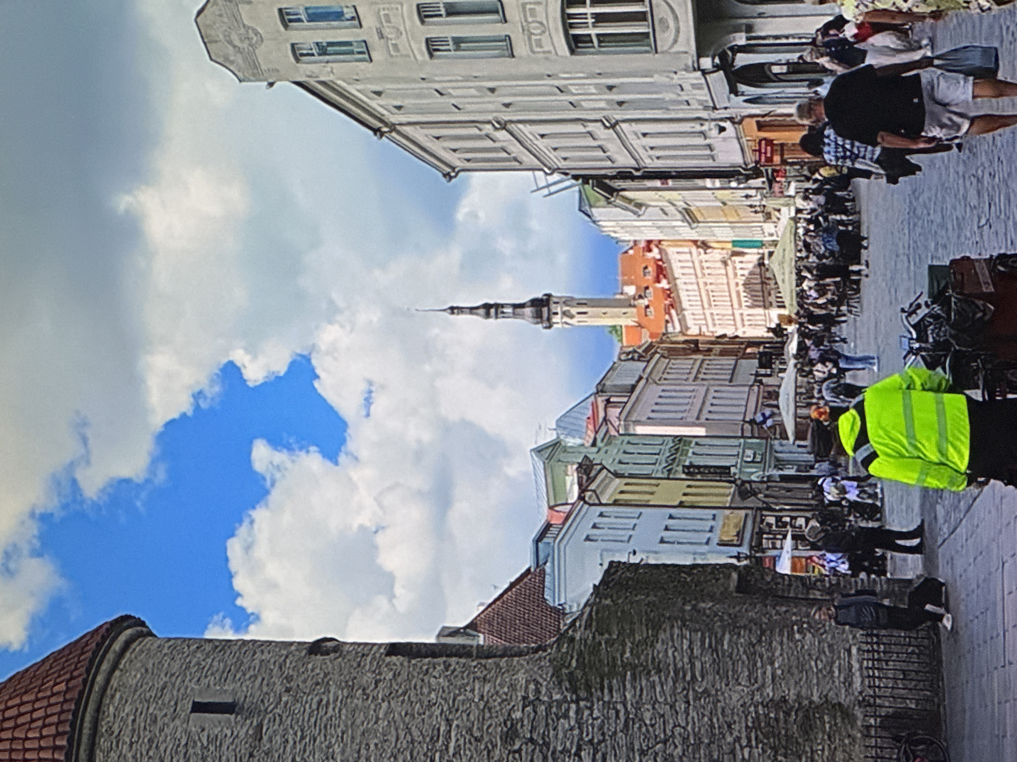 A vertical, eye-level, full shot depicts a bustling street scene in Tallinn, Estonia, under a partially cloudy blue sky. In the foreground left, a large, weathered stone tower with a red, conical roof juts into the frame. Its rough-hewn stone facade features a small, dark, rectangular window. A black metal fence runs along the base of the tower. Several people are visible near the tower: a person in a dark grey jacket and dark pants, pulling a black suitcase on wheels; another person in dark clothing; and further into the scene, a person wearing a bright, lime-green high-visibility jacket and dark pants, facing away from the camera.
The street is paved with cobblestones and lined with colorful, historic buildings on both sides. The buildings exhibit a variety of architectural styles and colors, including pale yellow, white, light blue, pink, and green facades, with red-tiled roofs. A prominent, slender church tower with a dark spire rises above the buildings in the mid-ground, centered in the perspective.
The street is populated with a crowd of people walking and milling about. Many are casually dressed. In the mid-ground, several outdoor seating areas with umbrellas are set up, suggesting cafes or restaurants. The overall atmosphere appears lively and commercial. The scene captures a moment of urban life in a historic European city.