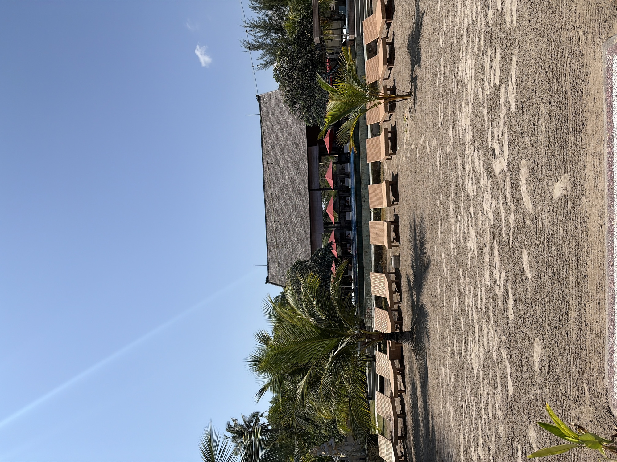 The image depicts a beach scene in Kampung Toyapakeh, Indonesia. In the foreground, a sandy beach is visible with numerous footprints and textured sand. Several rows of empty, light brown lounge chairs are arranged on the sand. A young palm tree stands near the center, with its fronds casting shadows on the ground. In the midground, a swimming pool is partially visible, bordered by a low wall. Behind the pool, a large building with a thatched roof is situated. Red umbrellas are open around the pool area, and there are trees and foliage surrounding the building. The sky is clear and blue, with a faint contrail visible on the left side. The overall atmosphere suggests a resort or hotel setting.