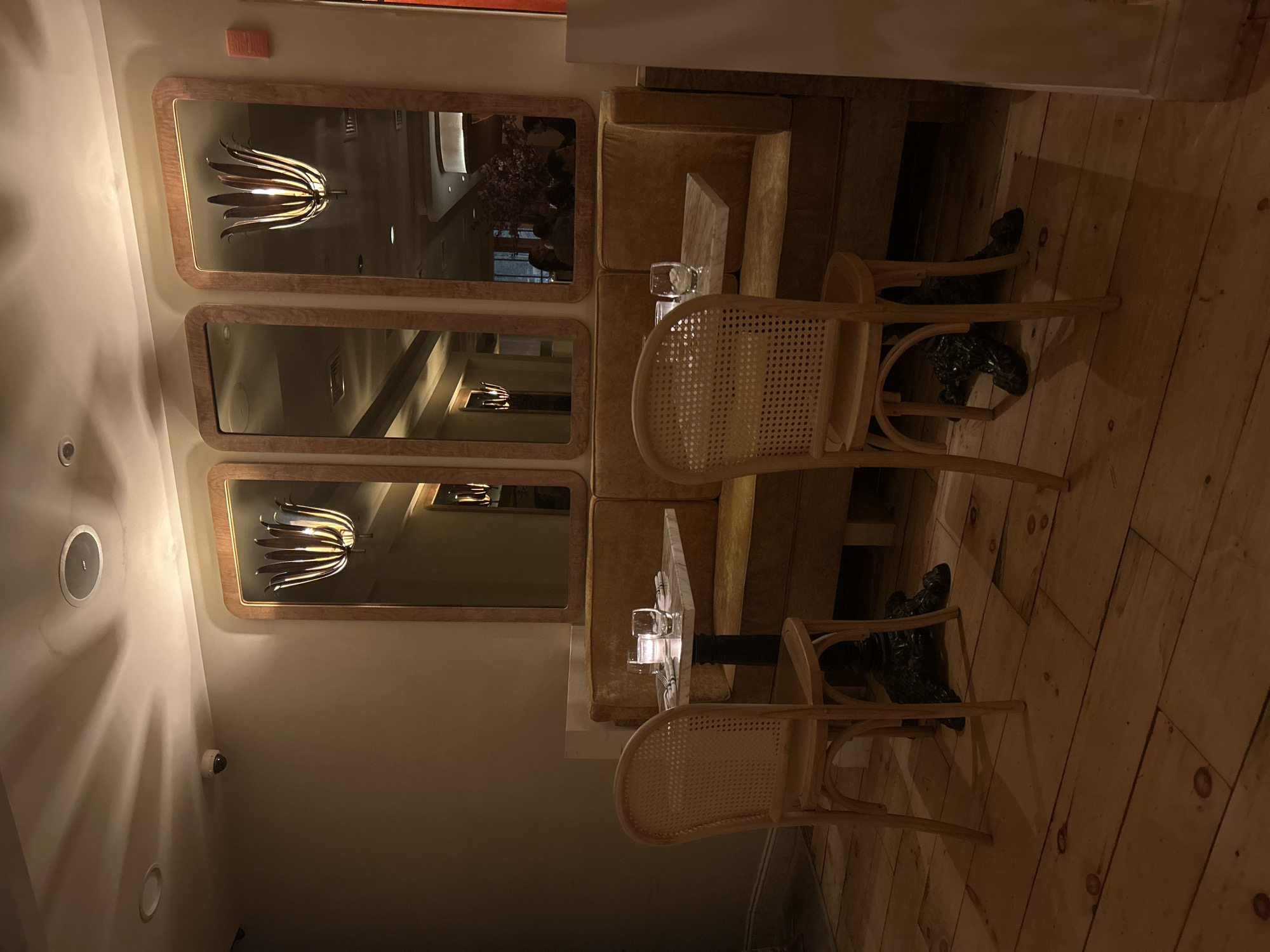 This image captures the interior of what appears to be a modern restaurant, cafe, or lounge, characterized by warm lighting and a mix of textures. The scene is dominated by a long, mustard-yellow upholstered banquette running along a wall, accompanied by several light-colored wooden chairs with woven cane backs and small, square-topped tables, possibly made of marble or a similar light stone.
On the wall above the banquette, three rectangular mirrors with wooden frames are vertically stacked. These mirrors reflect parts of the room, including a distinctive modern light fixture with multiple metallic or wooden "petals" radiating outwards, visible in the top and bottom mirrors. The middle mirror's reflection shows a blurred view that includes what might be a person's head or a large decorative floral arrangement, hinting at other patrons or decor in the space.
The floor is made of light-colored wooden planks. Underneath one of the chairs, a pair of black shoes or boots is visible, suggesting someone was recently seated or is still present just out of frame. The lighting is dim and atmospheric, with recessed lights in the ceiling casting soft glows. A small, white security camera is mounted on the wall. A small orange panel, likely a fire alarm, is also visible near the top left corner of the original image, but no text on it is legible. The room appears to be empty of active patrons, giving a quiet, inviting ambiance.