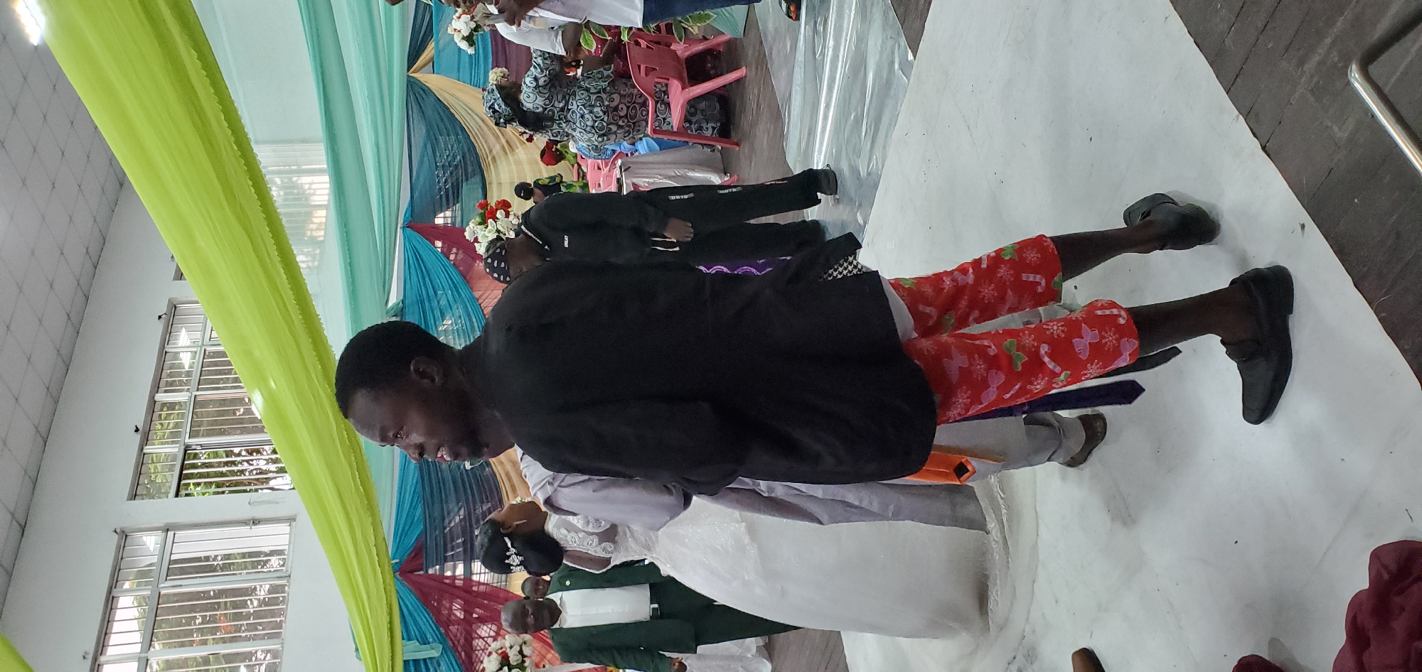 The scene captures what appears to be a wedding reception or event within a building in Bukuru, Nigeria. A couple is standing closely together, with the bride in a white gown and the groom in a black jacket and red Christmas-themed pants. They are surrounded by other people, including attendees seated in pink chairs, and others in darker clothing. The event is decorated with colorful fabrics draped as decorations. The floor is covered with a white fabric or covering. The presence of a window indicates it is an indoor setting.