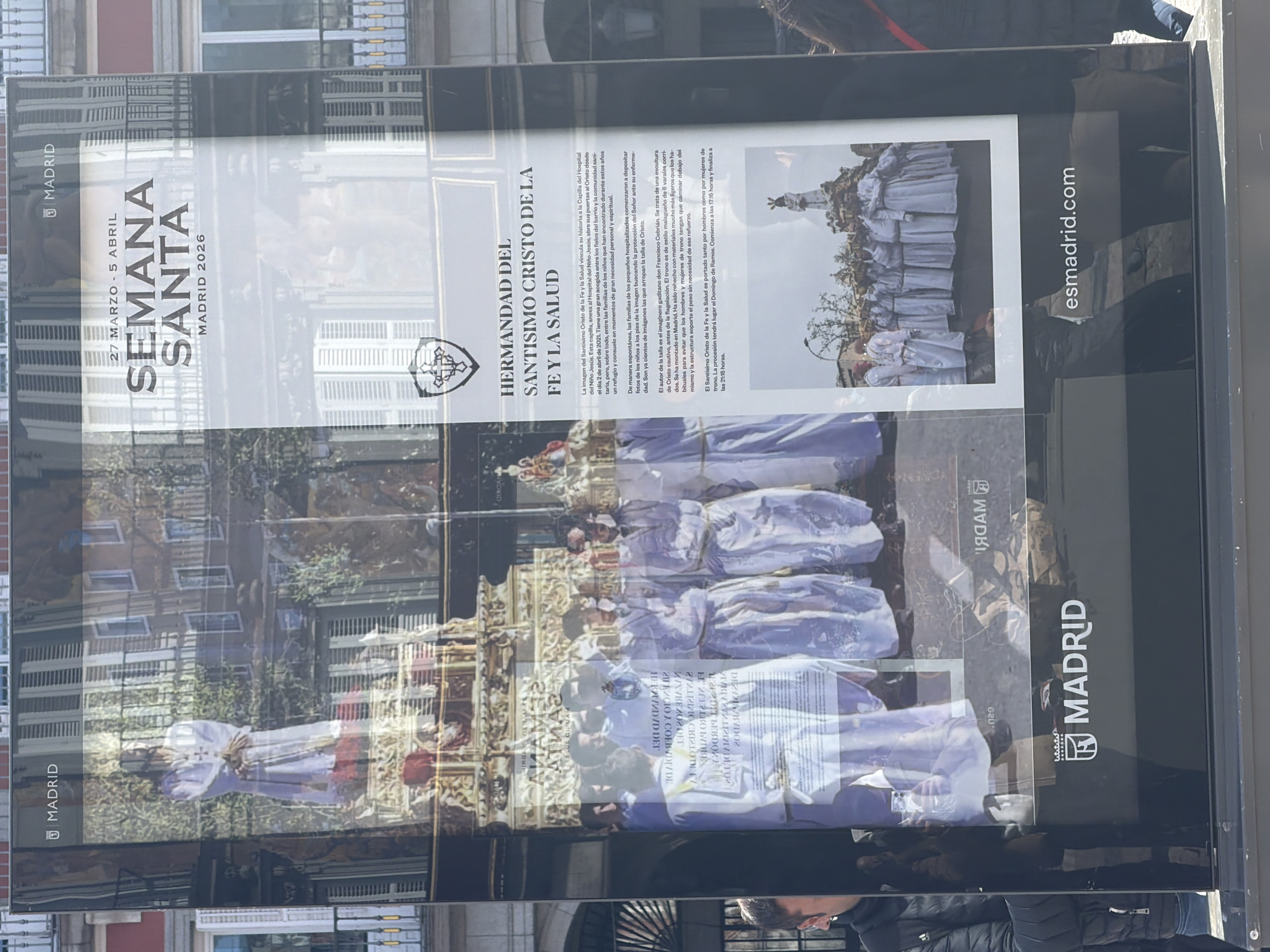 The image shows a promotional poster for the "Semana Santa" (Holy Week) in Madrid, Spain, scheduled for March 27 to April 5, 2026. The poster features the "Hermandad del Santisimo Cristo de la Fe y la Salud" (Brotherhood of the Holy Christ of Faith and Health). The poster displays an image of a religious procession with people carrying a statue of Christ. There is text describing the history and significance of the brotherhood and its connection to the Hospital del Niño Jesús (Children's Hospital). The poster includes the website "esmadrid.com" and the city name "Madrid" with the city's official logo. The poster is displayed in a public place, likely a street in Madrid, as reflections of buildings and people are visible on its surface.