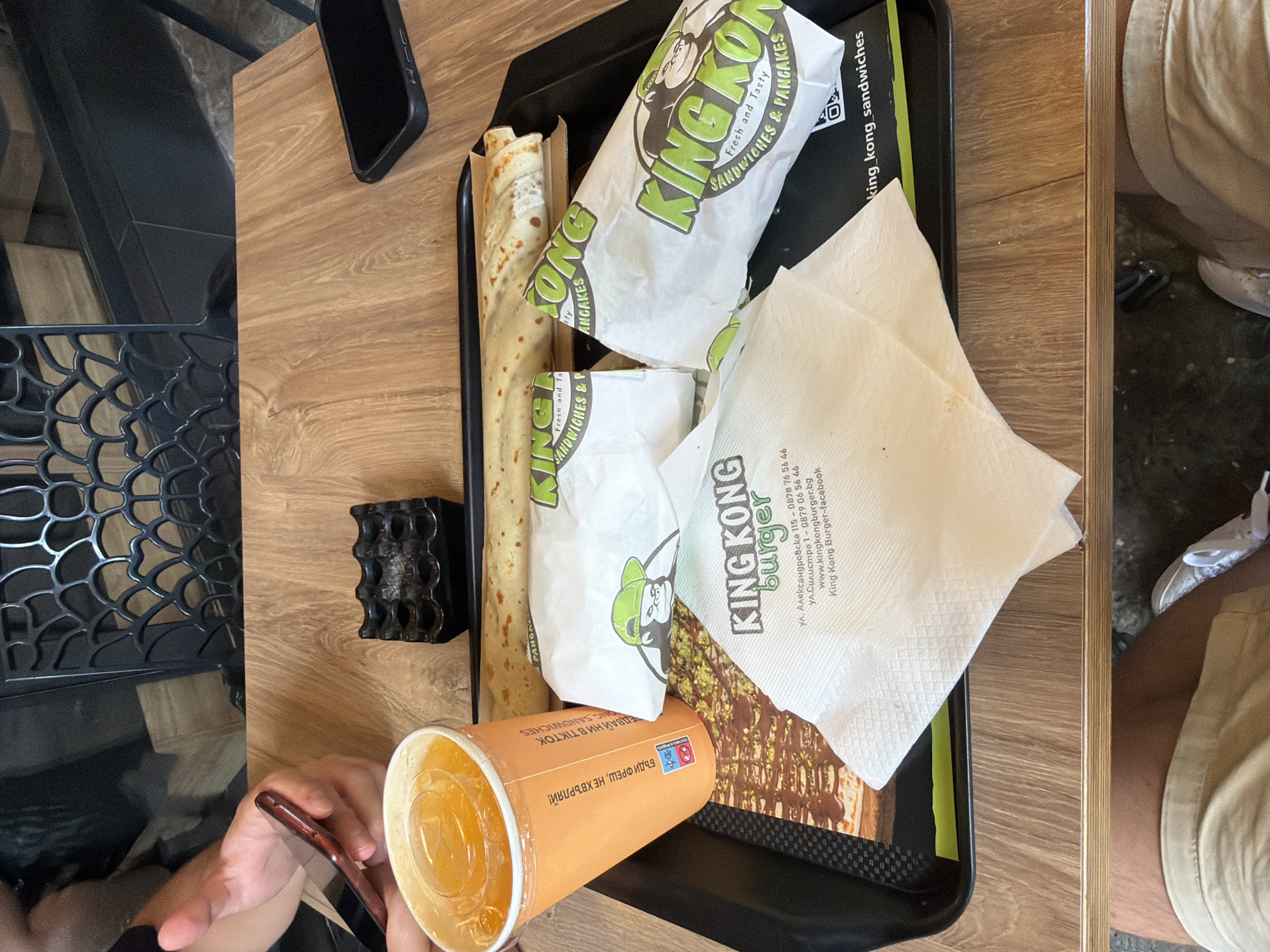An overhead shot captures a tray of food and drinks on a wooden table. The tray holds a large orange cup with a lid and straw, filled with an orange beverage, and two wrapped sandwiches with "King Kong Burger" branding. A rolled-up item, possibly a pancake or burrito, lies next to the sandwiches. A white napkin with "King Kong Burger" printed on it is placed on top of the wrapped items, showing contact information and a website for King Kong Burger. The napkin details address and phone number as: ul. Aleksandrovska 115 - 0878 76 56 46, www.kingkongburger.bg, King Kong Burger-facebook. This is the location of King Kong Burger in Burgas, Bulgaria. The scene appears to be indoors, with a black ashtray visible on the table. A person's hand holding a smartphone is partially visible in the upper left corner of the image. The overall impression is a casual dining setting.