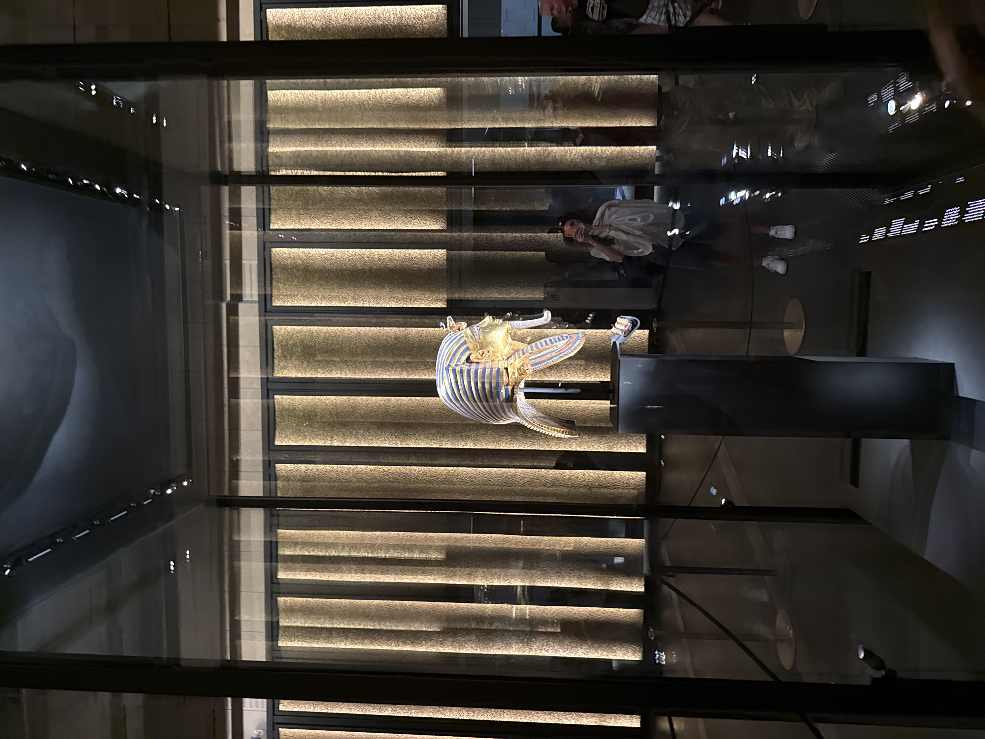An artifact, possibly a funerary mask, is displayed in a glass case. The artifact is gold and blue, with intricate patterns. It sits on a dark pedestal. Behind the display case, vertical panels of textured gold fabric are illuminated by vertical light fixtures, creating a striped effect. In the reflection on the glass, a woman is visible holding a phone, appearing to take a picture. She is wearing a white top and dark shorts. Another person, a man, is partially visible to the right, wearing a dark shirt and plaid shorts. The floor is dark and reflective, showing more reflections of the lighting. The setting appears to be a museum. The city of Kafrat Nassar, Egypt, is not depicted in this image.