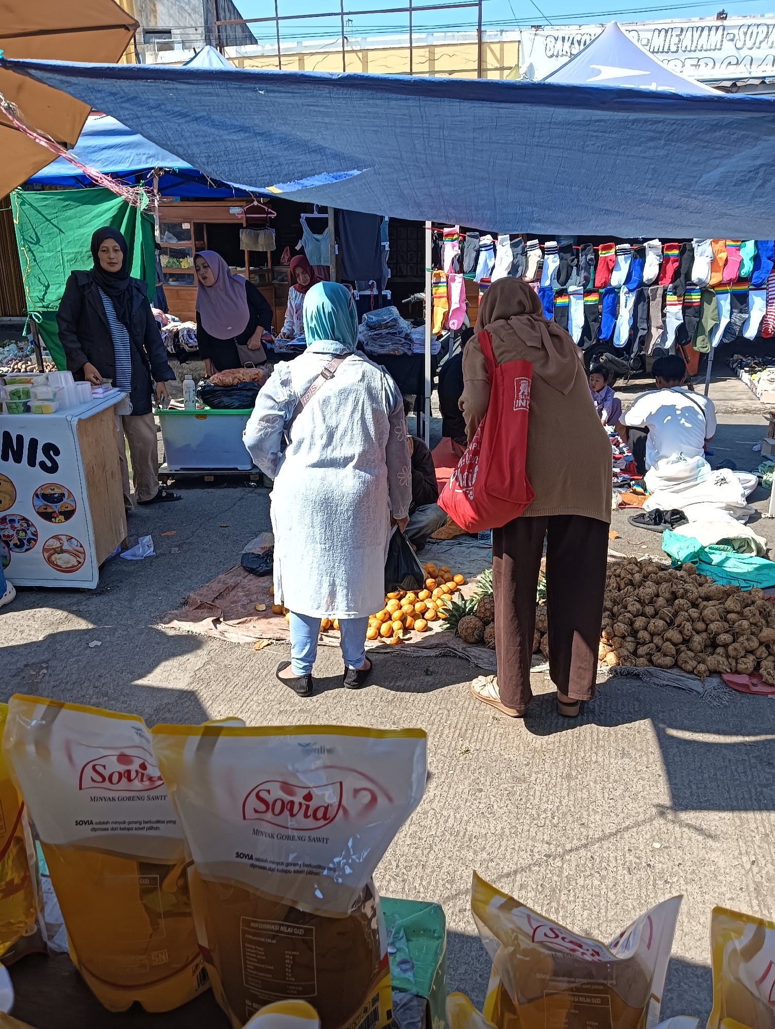 This is an outdoor market scene in Katapang, Indonesia, likely on a sunny day given the shadows. The image captures a bustling commercial environment with several people engaged in transactions or browsing.
In the foreground, there are two large plastic pouches of "Sovia Minyak Goreng Sawit" (cooking oil), with nutritional information visible on one. To the left, a stall displays white containers with yellow liquid, possibly food items, and signage that includes the word "NIS".
Further into the market, multiple individuals are present. A woman in a black jacket and headscarf stands behind a stall. Another woman in a light purple headscarf is interacting with her. In the mid-ground, a person wearing a light blue hijab and white tunic is facing away from the camera, seemingly examining goods. To the right of this person, a woman in a brown hooded sweater and dark pants, carrying a red tote bag, is looking down. A child in a purple outfit is visible in the background.
The market features various goods on display. Rows of colorful socks are hung under a blue tarp. To the right, a large pile of root vegetables, possibly ginger or similar produce, is displayed on a mat. Scattered around the area are also some fruits, including oranges and pineapples.
The environment is typical of an Indonesian street market, with temporary stalls, tarps providing shade, and a paved ground surface. The background shows buildings and a large white banner advertising "BAKSO - MIE AYAM - SOP KAKI SAPI". The overall impression is one of active commerce and daily life in a local Indonesian community.