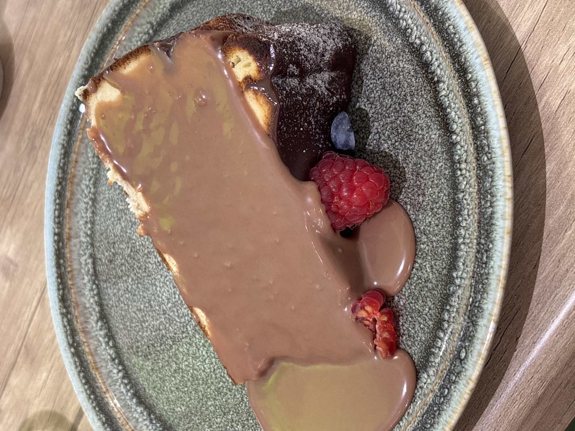 A close-up, angled shot of a piece of dessert on a decorative plate placed on a wooden surface. The dessert appears to be a slice of cake, possibly a layered sponge cake. The cake is topped with a generous pour of smooth, light brown chocolate sauce, obscuring much of the cake's surface. Part of the cake shows a darker, possibly chocolate-covered layer dusted with a white powder, likely powdered sugar. The plate is adorned with several fresh berries, including a vibrant red raspberry and a single blueberry. Two small, reddish berry fragments are also present, partially covered by the chocolate sauce. The plate has a distinct texture and color pattern, with a mix of gray, green, and brown shades, and a somewhat uneven, textured surface. The wooden surface beneath the plate has a light brown, slightly glossy appearance, suggesting it might be a table or countertop. The specific location of the image is Unknown, Unknown.