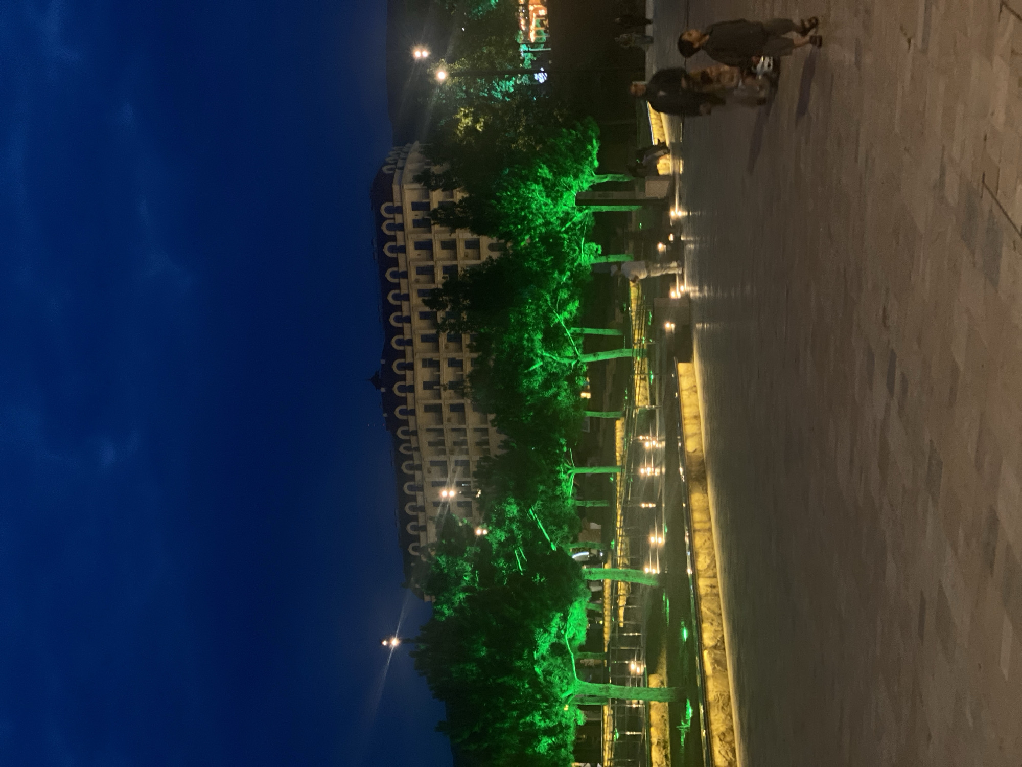 The image depicts a street scene at night in Derbent, Russia. A grand building with arched windows is illuminated, lined with trees that are bathed in vibrant green light. The street or walkway in the foreground is paved with large, light-colored stones, and is wet, reflecting the lights from the scene.
Several people are visible walking on the paved area. One person is walking towards the viewer, and another is walking away. Further in the background, more figures can be seen.
The sky is a deep, dark blue, indicating it is nighttime. The overall atmosphere is illuminated by artificial lighting, creating a striking contrast between the dark sky and the brightly lit building, trees, and walkway. The wet pavement suggests recent rain or a deliberate watering of the area. The scene appears to be a public space, possibly a promenade or a street in front of a hotel or a significant building, given its architectural style. The green lighting on the trees adds a festive or decorative touch to the environment. No visible text is present in the image.