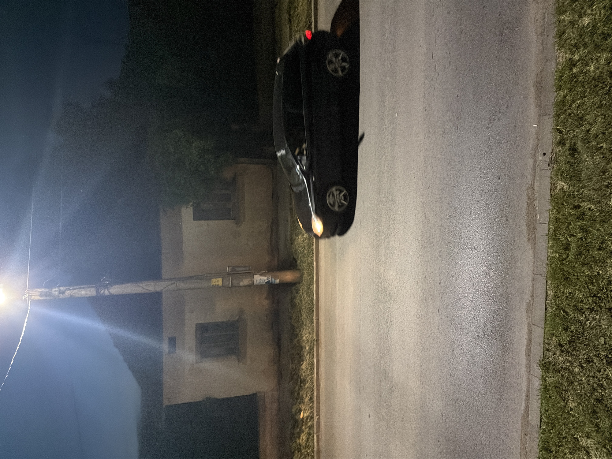 The image depicts a dark-colored sedan parked on the side of a street at night, likely in Iskar, Bulgaria. The photograph is oriented vertically, showing the car on the right half of the frame with its front facing the bottom and rear towards the top (when correctly oriented). The car's headlights are illuminated at the front, and a red taillight is visible at the rear. Its wheels feature a multi-spoke design.
To the left of the car, an asphalt road stretches across the frame, bordered by a concrete curb and a grassy verge on its left edge. Beyond the verge, a brightly lit street lamp on a pole illuminates the scene, casting a strong light flare. Directly behind the lamp post, a light-colored building with multiple rectangular windows is visible in the background. One window appears to have horizontal slats or bars. Above the building, the dark outlines of trees or dense foliage are visible against the night sky.
Several utility wires and small boxes are attached to the street lamp's pole, some extending towards the building. Faint, illegible text or numbers are visible on a small white or light-colored label or sign affixed to the pole structure, including what might be "Ape" or "Are" and numbers like "230". The overall setting suggests a quiet urban or suburban street scene under artificial light at night. No people are visible.