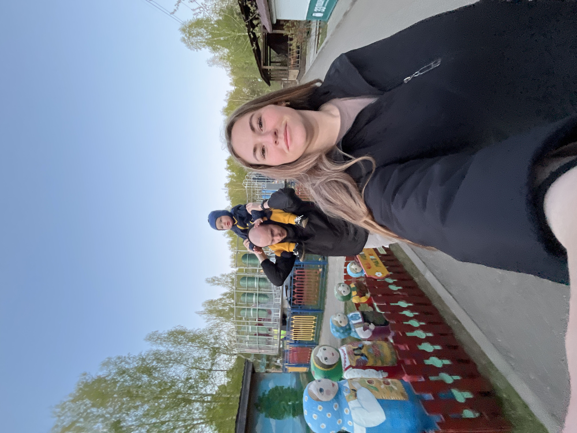 A selfie taken outdoors features a woman with long, light brown hair. She is wearing a black jacket. Behind her is a man with a child on his shoulders. The man is wearing a black jacket and the child has a blue hat. Further in the background is a playground or park area with a colorful fence and what appears to be wooden dolls. The location is Chaikovsky, Russia.