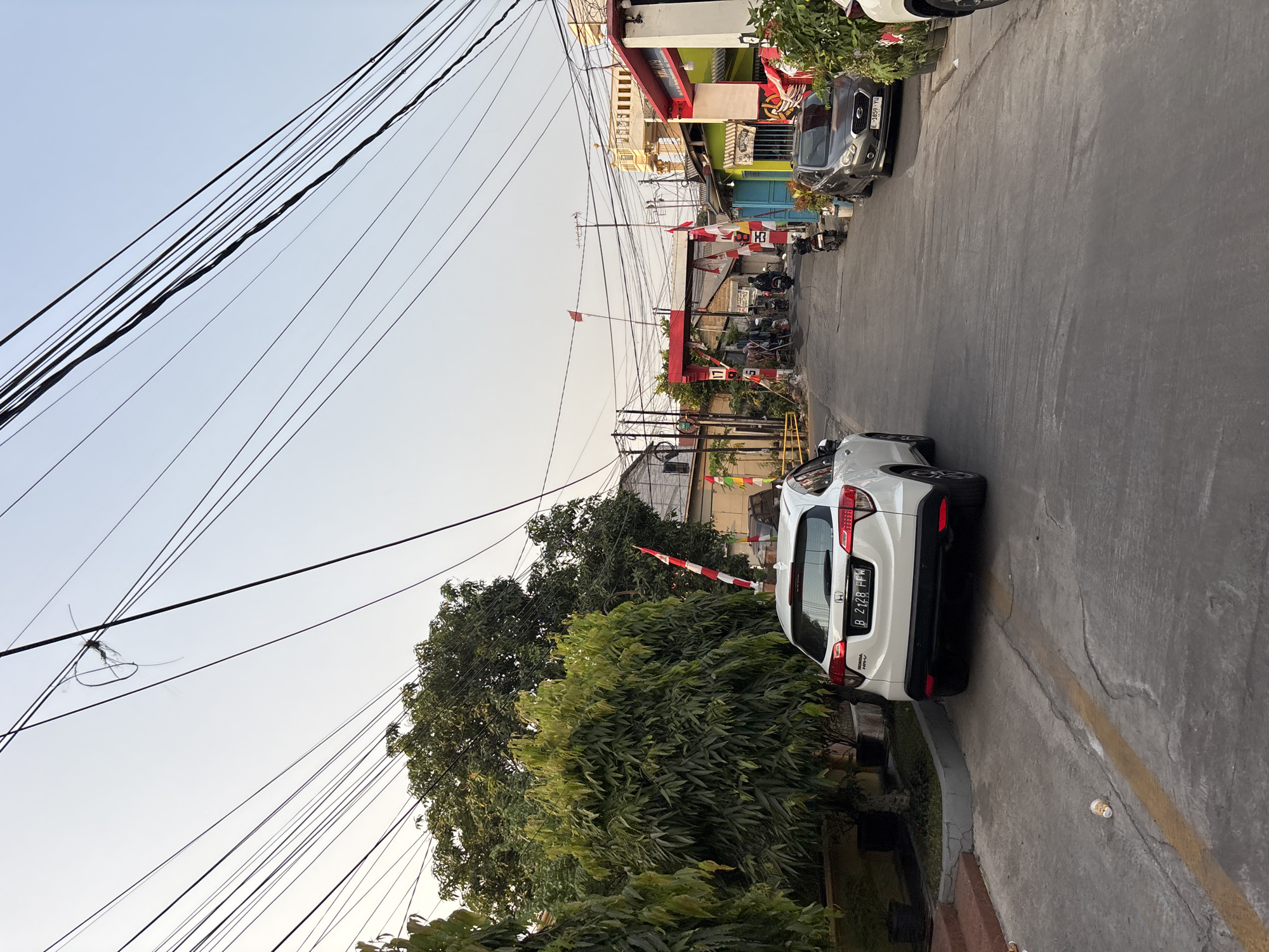 A white Honda HR-V is parked on the left side of a narrow, paved road in a residential area of Bekasi, Indonesia, with its rear facing the camera. The car's license plate reads "B 2128 FFW". A grey Datsun GO is parked on the right side of the road further down, its license plate reading "E 1859 YU". Overhead, a dense network of utility wires crisscrosses the sky. The road is lined with trees and houses, some of which are painted in bright colors. A red and white archway, decorated with flags, spans the road in the distance, with several motorcycles and people passing underneath. The overall scene depicts a typical day in a bustling Indonesian neighborhood.