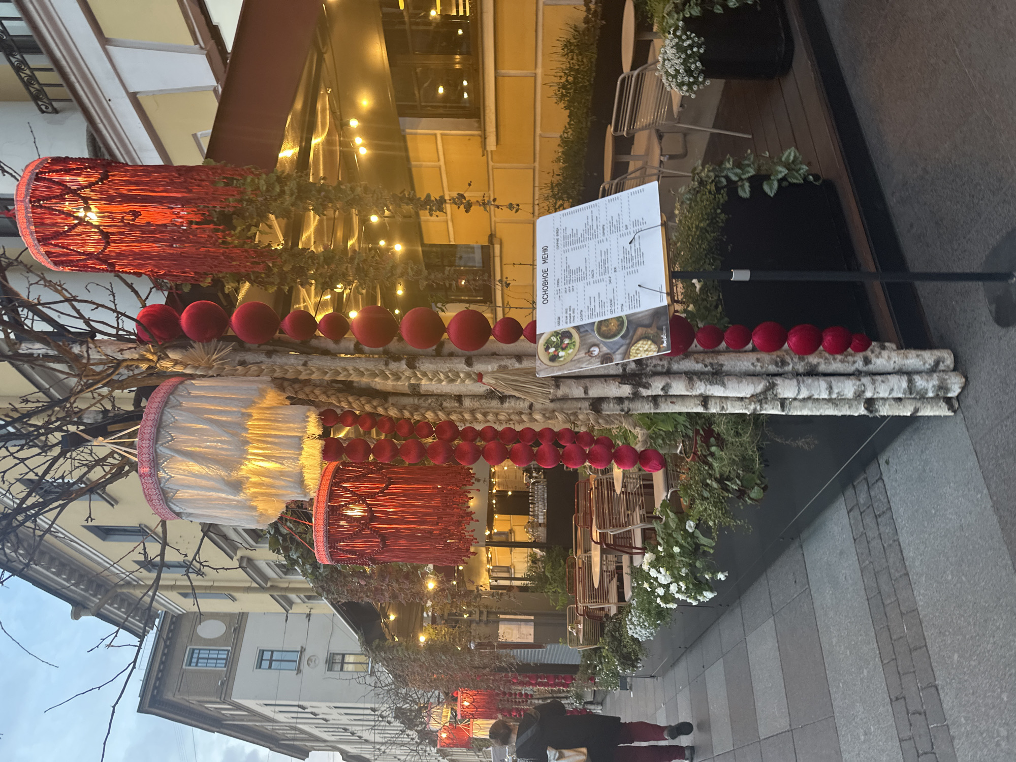 The image depicts an outdoor cafe setting in Saint Petersburg, Russia. Red and white cylindrical lanterns with intricate designs hang from bare branches, creating a festive atmosphere. A string of red spherical ornaments adorns a horizontal arrangement of birch logs and braided material, likely serving as a decorative barrier or display. A menu board displaying the restaurant's offerings stands upright behind the decorative arrangement. Tables and chairs are visible in the background, suggesting a dining area. Pedestrians are walking along the sidewalk in the lower part of the image. The architectural style of the buildings suggests the location is within the historical city center.