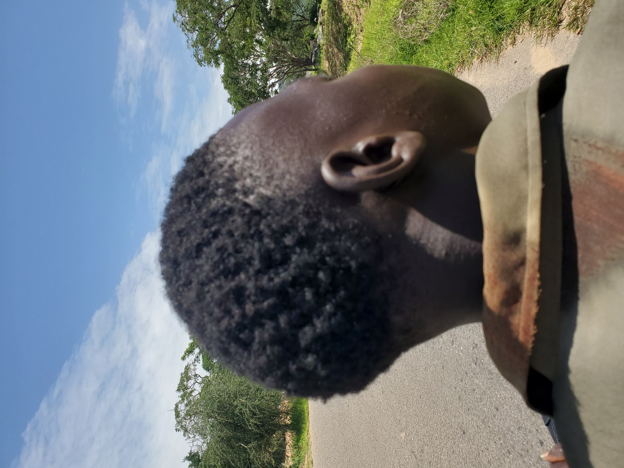 A close-up, over-the-shoulder shot shows the back of a young person's head, with dark, short, textured hair. The person is wearing a khaki collared shirt with a weathered, possibly rusted, band around the neck area. The person is facing away from the camera, looking towards the right of the frame.
The background features a paved road stretching into the distance, bordered by a grassy verge and various trees. The sky above is a vibrant blue with scattered white clouds, suggesting a sunny day. The scene appears to be in a rural or semi-rural outdoor environment. There is no specific information in the image to indicate the location is Gwolong, Nigeria.