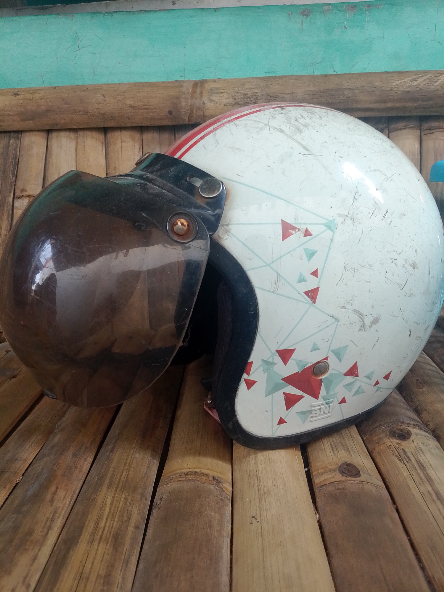 A white motorcycle helmet with a brown visor rests on a surface of weathered bamboo planks. The helmet has red and light blue geometric patterns and is marked with visible scratches and scuffs. It is positioned on the right side of the frame. The background features a turquoise wall with vertical bamboo slats underneath, suggesting an outdoor or rustic setting. The overall impression is one of a casually placed item in a natural, weathered environment. The scene appears to be located in Tasikmalaya, Indonesia, as indicated by the common use of bamboo for construction and decorative elements in the region.