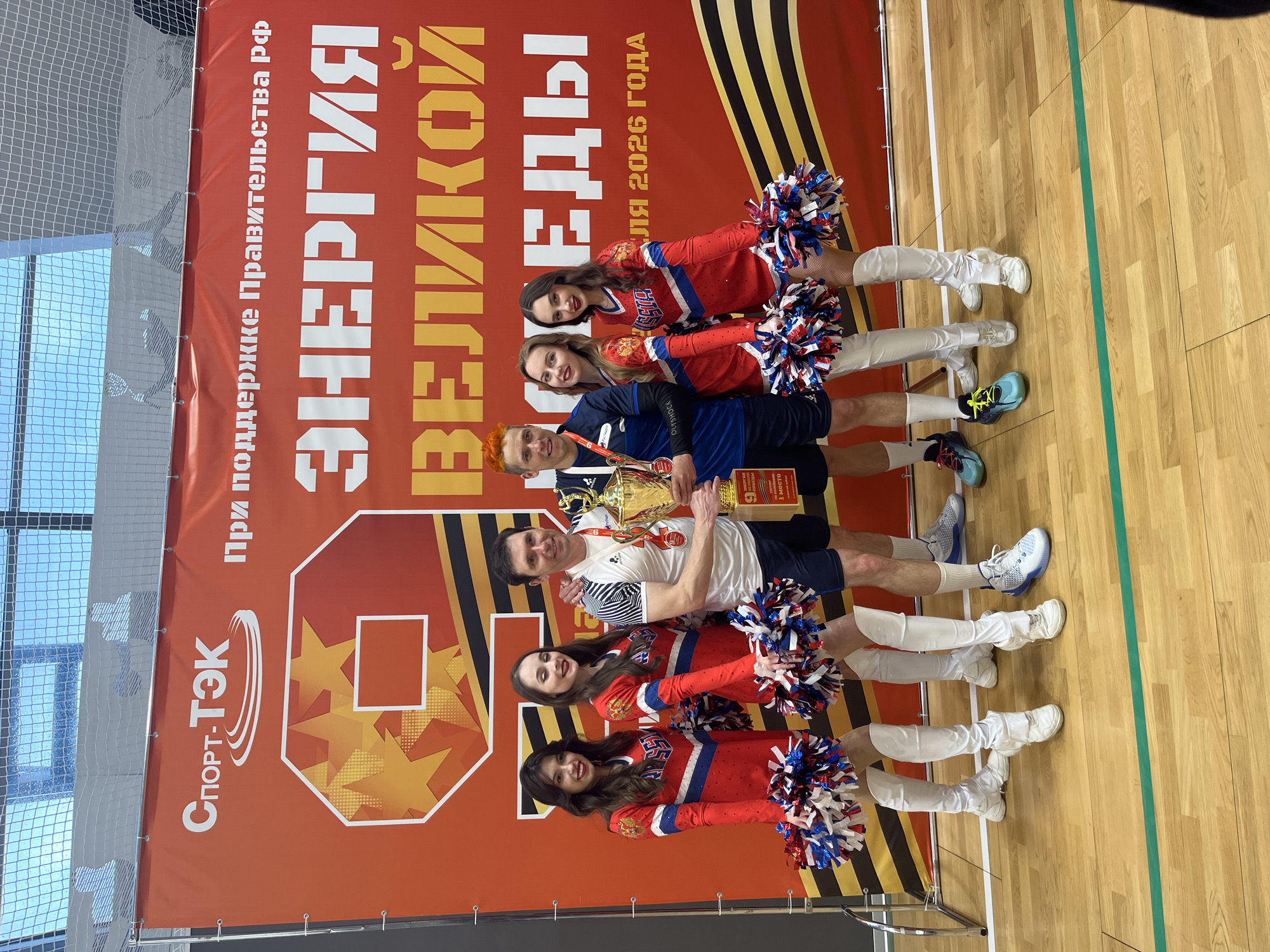 The image shows a group of people posing in front of a banner. The banner features the logo of "Спорт-ТЭК," and text in Russian reading "При поддержке Правительства рф ЭНЕРГИЯ ВЕЛИКОЙ ВОВЕДЫ ля 2026 ГОДА" which roughly translates to "With the support of the Government of the Russian Federation, Energy of the Great Vovods for 2026." There are at least four people in athletic attire, standing in the middle of the group and holding a trophy. Flanking them are four cheerleaders, two on each side, holding pom-poms. The floor beneath them is made of polished wood. The photo appears to have been taken indoors, possibly in a gymnasium or sports arena. The location appears to be in Moscow, Russia based on the Russian Cyrillic writing on the banner.