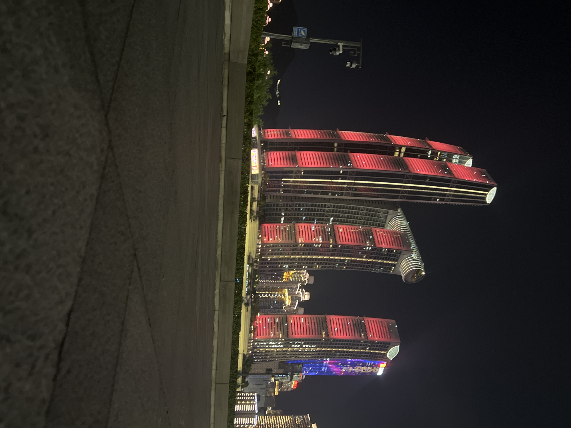In a nighttime city scene in Jiangbeicheng Subdistrict, China, several illuminated skyscrapers dominate the background. The most prominent structures are tall, modern buildings with striking red and yellow lighting. One building on the left features vertical red lighting panels interspersed with yellow vertical lights, capped with a circular illuminated element. To its right, another skyscraper has a curved, top section with circular lighting and red vertical panels. Further to the right, a third building displays vibrant red and blue digital lights. In the foreground, a dark asphalt road or plaza with visible grid lines stretches towards the viewer. A concrete barrier with green bushes and trees behind it separates the foreground from the buildings. To the left, a lamp post with two surveillance cameras and a sign is visible. The sky is dark and clear, indicating nighttime.