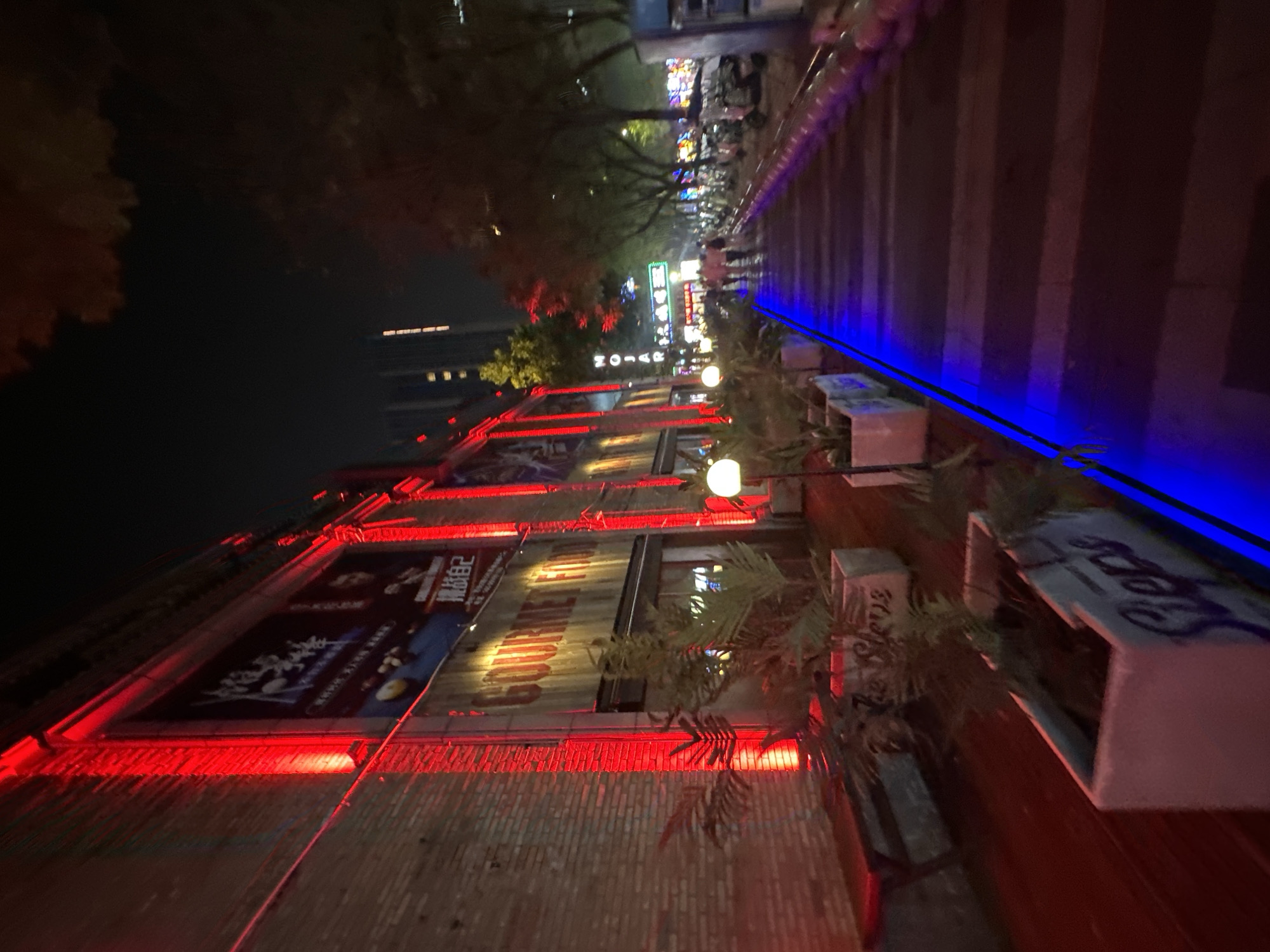 The image depicts a nighttime scene on a street in the Chongqing High-tech Zone, China. The left side of the image features a brick building facade illuminated by vibrant red lights, with banners and signage visible. To the right, a pathway is lined with plants in planters and illuminated by blue lights along its edge. The pathway recedes into the distance where it merges with a street featuring more lights, parked motorcycles, and a few pedestrians walking. The overall atmosphere is that of a commercial or entertainment district at night.