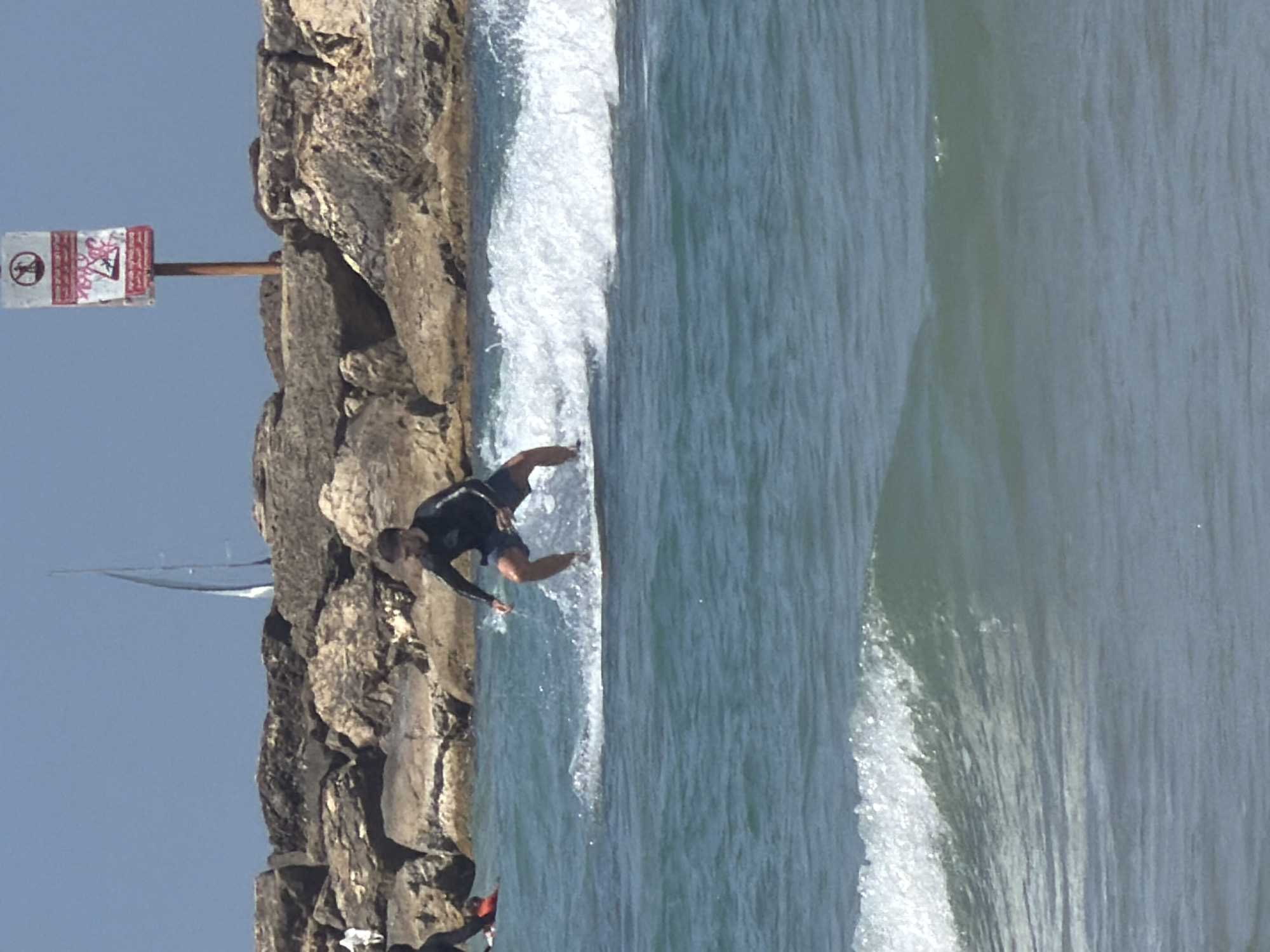 A man in a wetsuit is pictured surfing a wave near a rocky breakwater. The image appears to be taken at a beach in Tel-Aviv, Israel, as evidenced by the signage warning against diving and the presence of a sailboat in the background. The water is choppy and the sky is clear. Another person can be seen in the lower left.