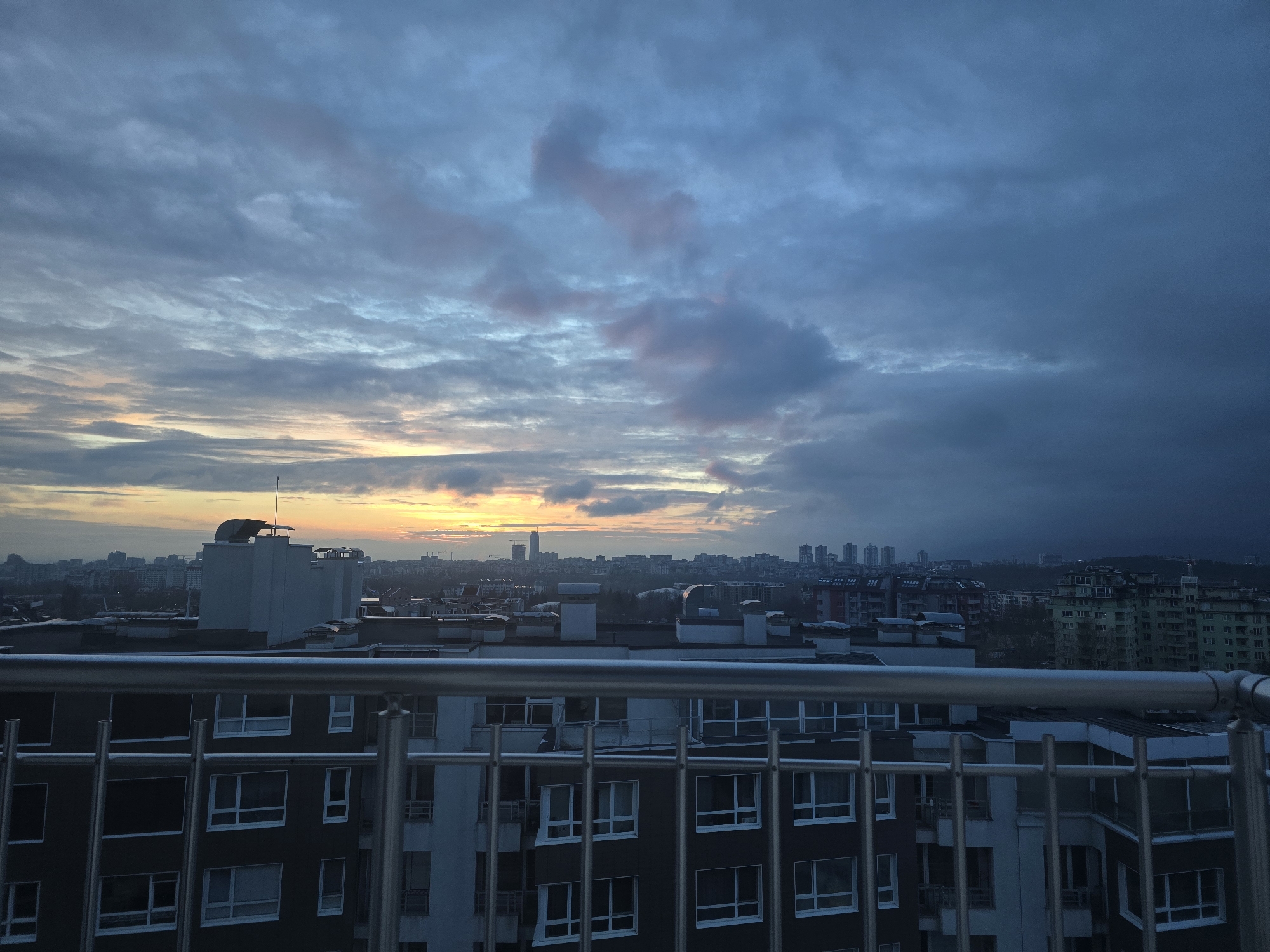 The image depicts a cityscape at dusk or dawn, viewed from an elevated position, likely a balcony. A metal railing is visible in the foreground. The cityscape appears to be Sofia, Bulgaria, with a mix of residential and commercial buildings. In the distance, a cluster of high-rise buildings can be seen, suggesting a central business district. The sky is filled with clouds, exhibiting hues of blue, grey, and a faint orange glow near the horizon, indicating either sunrise or sunset. The overall ambiance is subdued and atmospheric.