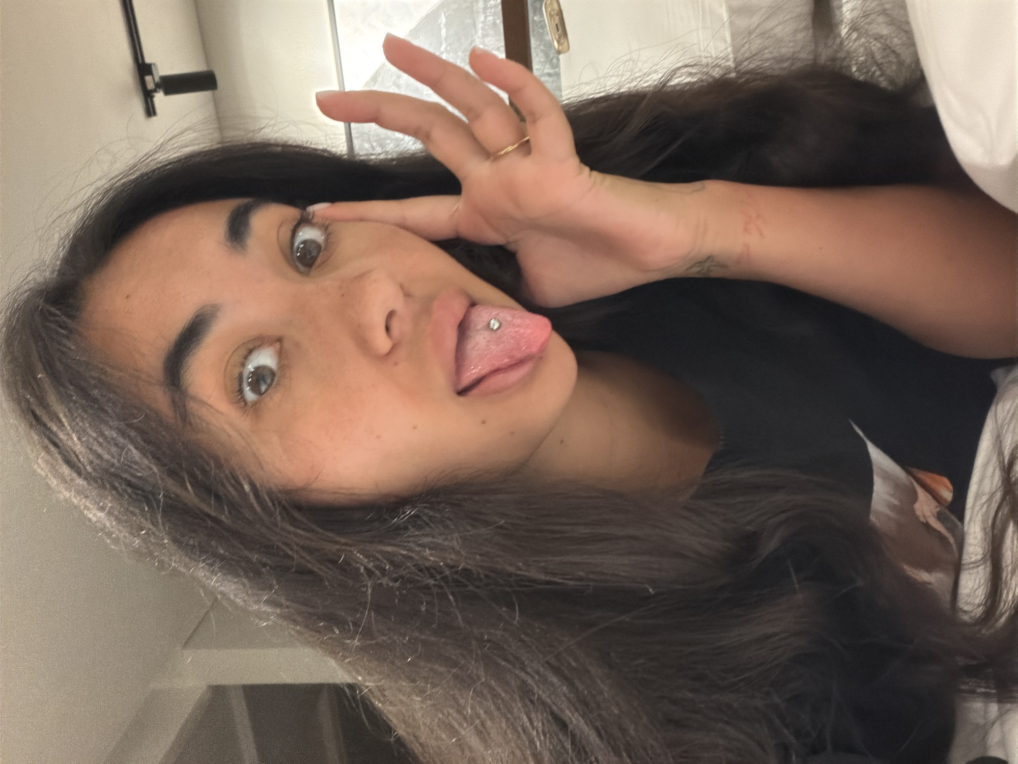 A young woman with long dark hair is looking directly at the camera. Her face is partially obscured by her hair, but her eyes and mouth are clearly visible. She is sticking her tongue out, which has a stud piercing in the center. Her fingers are slightly curled and positioned near her eye, perhaps as a gesture. A gold ring is visible on one of her fingers. The background appears to be an indoor setting, likely a bedroom or hotel room, given the presence of a white pillow in the upper right corner and a wall in the upper left. No specific location details relating to Tibubeneng, Indonesia are evident in the image.