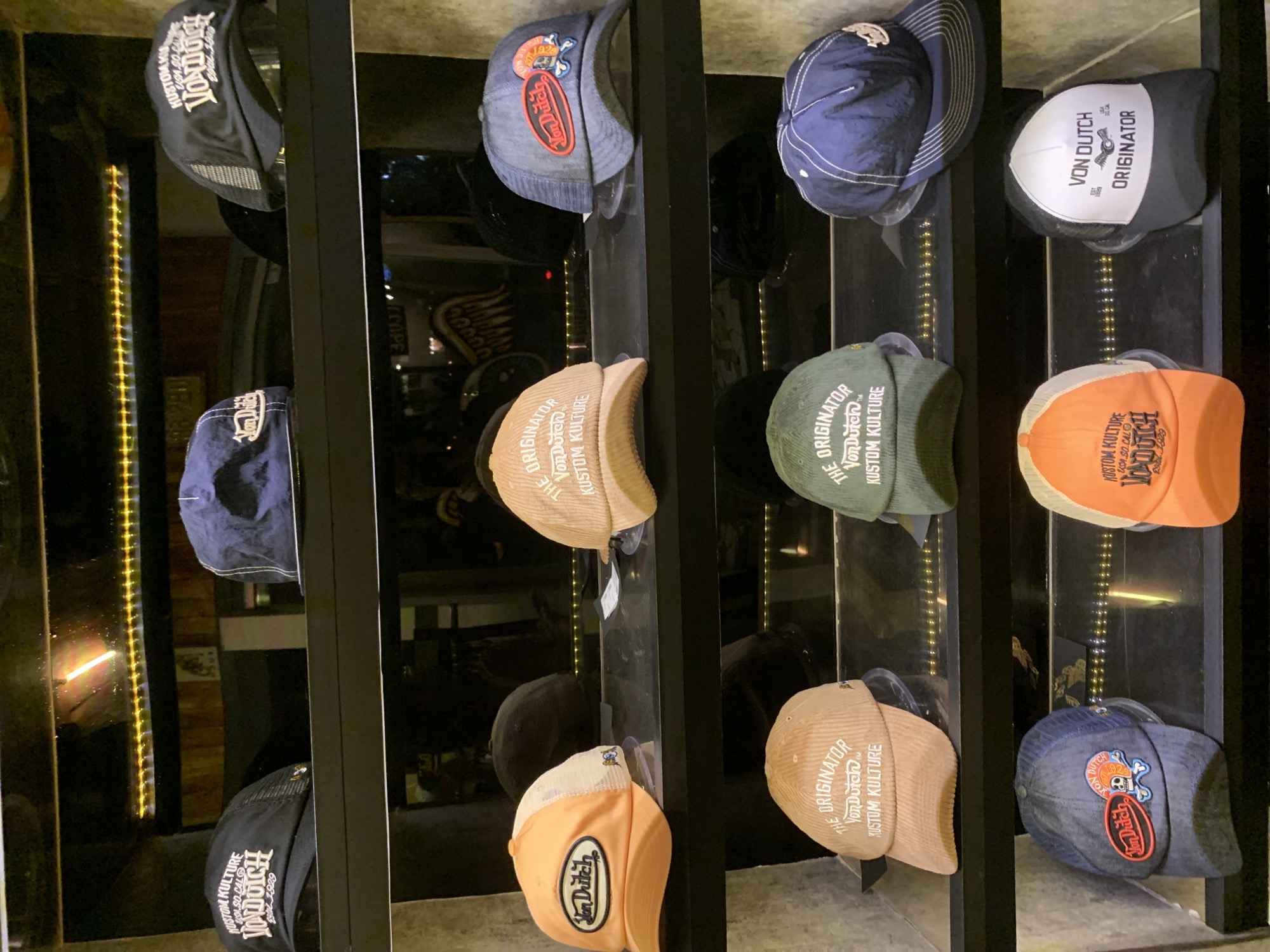 The image depicts a display of Von Dutch hats in what appears to be a retail setting. The hats are arranged in rows on a black metal frame, presumably inside a store in Ubud District, Indonesia. The hats vary in color and design, with some featuring the "Von Dutch" logo prominently, others displaying "The Originator" and "Kustom Kulture" branding. Backlighting is present, illuminating the merchandise and creating a visually appealing display. There are no people visible in the frame. The setting implies a commercial environment focused on fashion accessories.