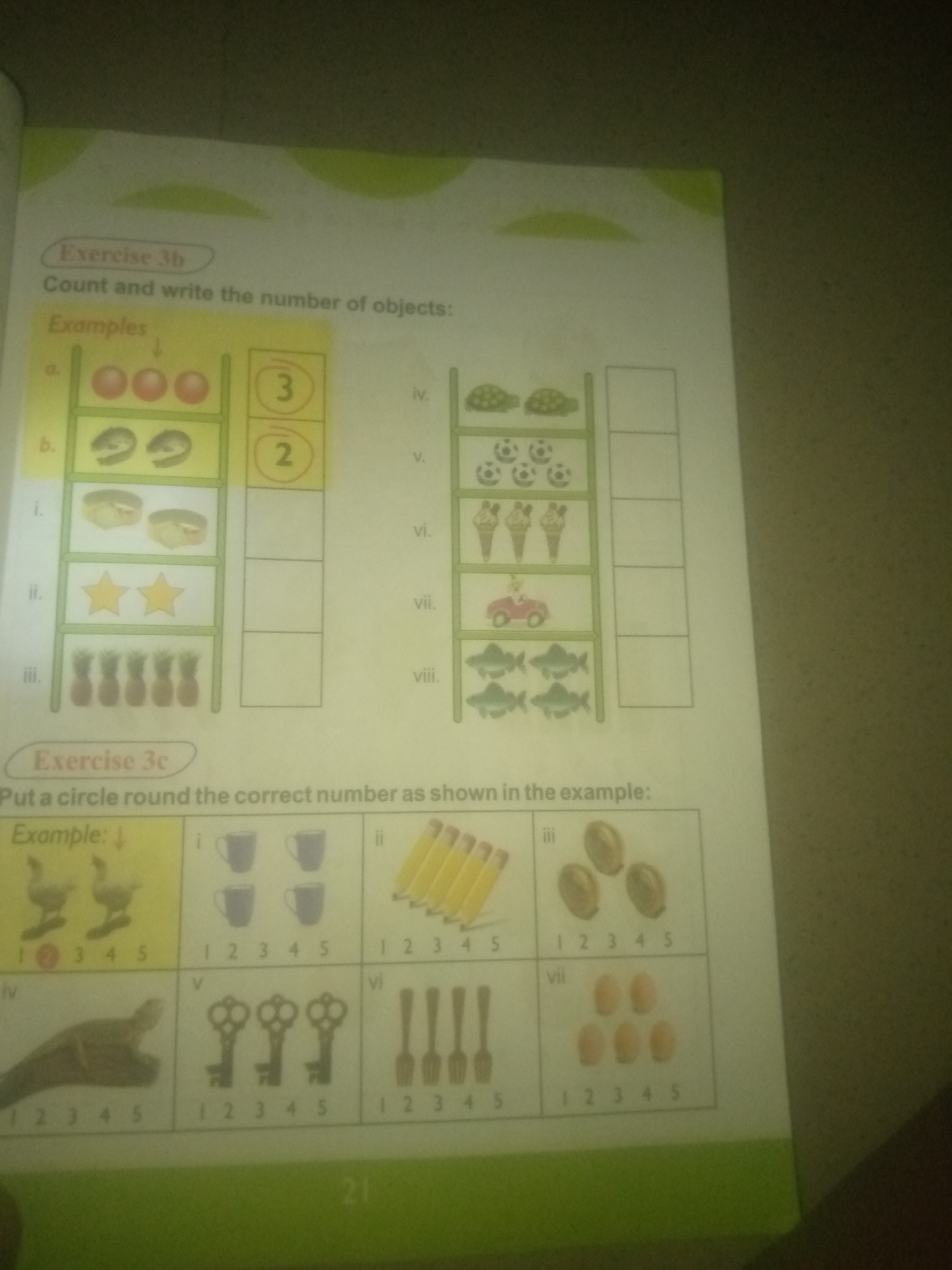 The image is a photograph of a page from a children's workbook, likely for early elementary education. The page contains exercises related to counting and number recognition.
The top section of the page is labeled "Exercise 3b" and instructs the user to "Count and write the number of objects." This section features a vertical ladder-like structure with different sets of objects depicted on each rung. To the right of these objects are numbered boxes, with some numbers already circled as examples. The objects shown include red balls, what appear to be fish or shrimp, pastries, stars, and pineapples.
The bottom section of the page is labeled "Exercise 3c" and instructs the user to "Put a circle round the correct number as shown in the example." This section presents a grid of smaller images, each with a series of numbers below it. The example shows two ducks, with the number '2' circled. Subsequent images in this section include cups, pencils, coins, keys, forks, and eggs.
The overall environment appears to be indoors, with the page lying on a surface that is not fully visible but has a textured, possibly carpeted, appearance. The lighting is somewhat dim, with a slight glare on the page, suggesting the photograph was taken with a flash or under artificial light. The image is cropped, and the bottom right corner shows a portion of a green border, typical of workbook pages. There are no people visible in the image.
The text on the page is in English. There is no information within the image that connects it to Jauro Yinu, Nigeria, or any specific city.
