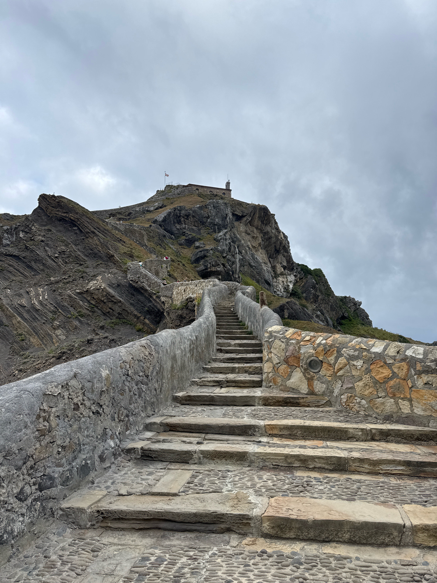 The image depicts a stone staircase ascending a rocky hillside towards a building topped with a flag, located at San Juan de Gaztelugatxe, an islet connected to the mainland by a bridge, near the town of Bakio, Spain. The sky is overcast with grey clouds. The staircase is constructed from rough-hewn stone steps and a textured cobblestone path, bordered by a weathered stone wall. Several individuals are visible ascending the stairs. The rocky terrain is layered with distinct geological formations, and sparse vegetation grows on the slopes. A small building with a tower and a flag is situated at the summit of the hill, with a railing in front of it.