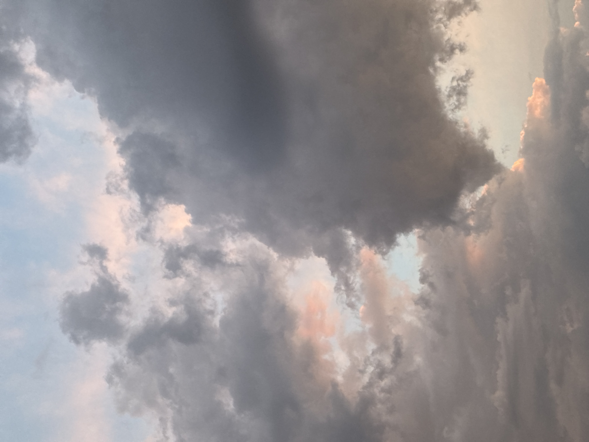 The image shows a close-up view of a cloudy sky. The clouds are a mix of dark gray and lighter, fluffy formations, with hints of pink and peach hues suggesting the presence of sunset or sunrise light. Patches of pale blue sky are visible through the gaps in the clouds. The overall impression is one of dramatic and atmospheric cloud formations. There are no discernible elements of a city, people, or specific objects within the frame.