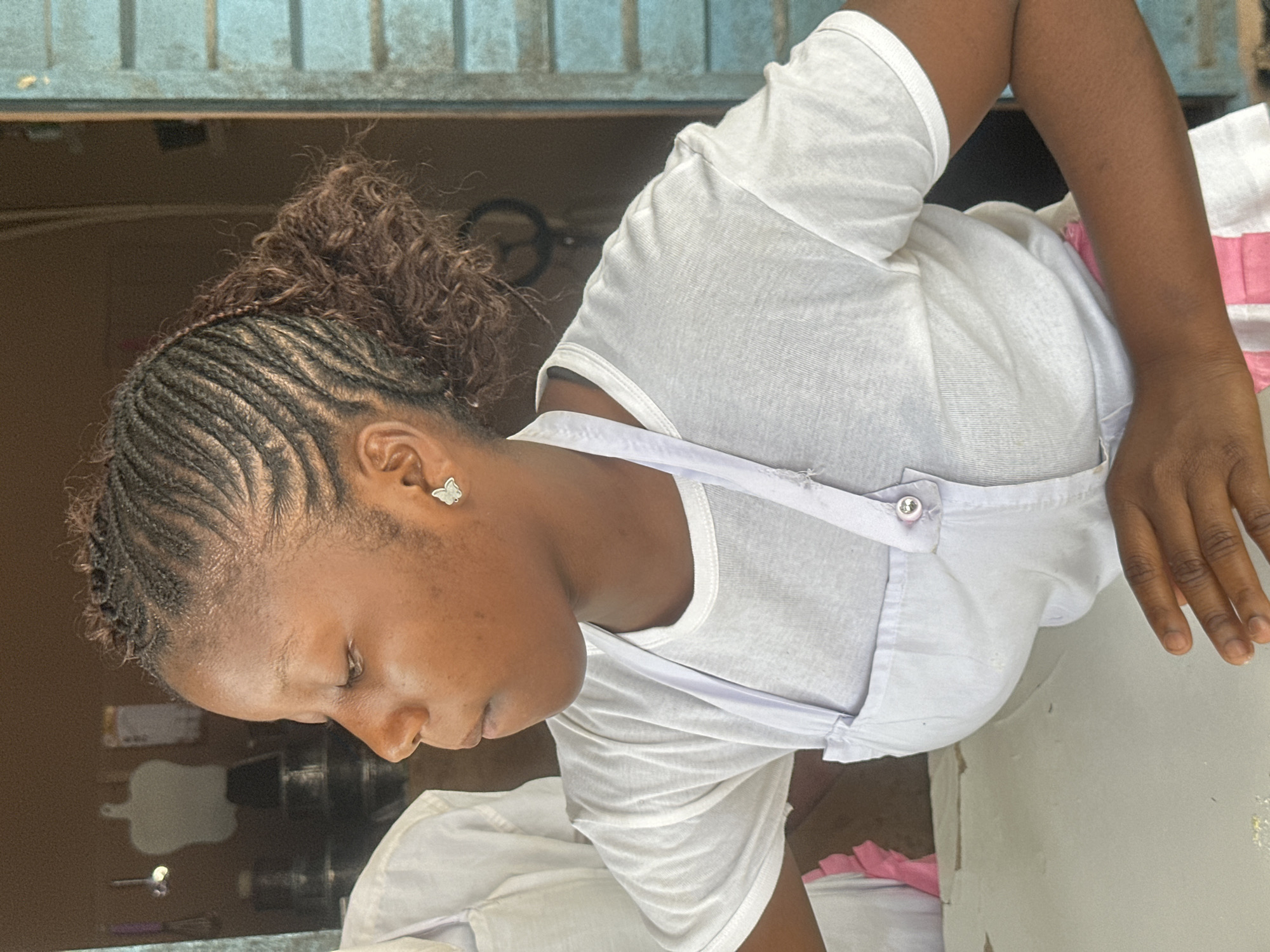 The image captures a young Nigerian woman in what appears to be a commercial setting in Badagry, Nigeria. She is wearing a white t-shirt and a white apron, with a pink garment visible underneath the apron. Her hair is styled in cornrows that are pulled back into a ponytail, and she is wearing a small butterfly-shaped earring. Her gaze is directed downwards and to her left, with her right hand resting on a light-colored surface. The background suggests an indoor space, possibly a shop or food stall, with shelves and items visible. The overall impression is of a working individual in an everyday urban environment.