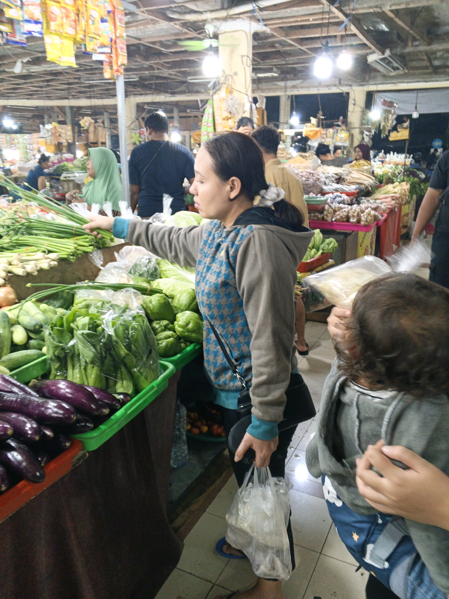 A woman in a grey hoodie with a blue patterned shirt underneath, holding a plastic bag of groceries, stands at a market stall in Denpasar, Indonesia. She is reaching for a bunch of green onions. In front of her are bins of vegetables, including eggplants and bell peppers. To her right, a child in a grey hoodie and blue pants looks at the groceries. In the background, other shoppers and vendors are visible. The market appears to be indoors with a high ceiling and fluorescent lighting.