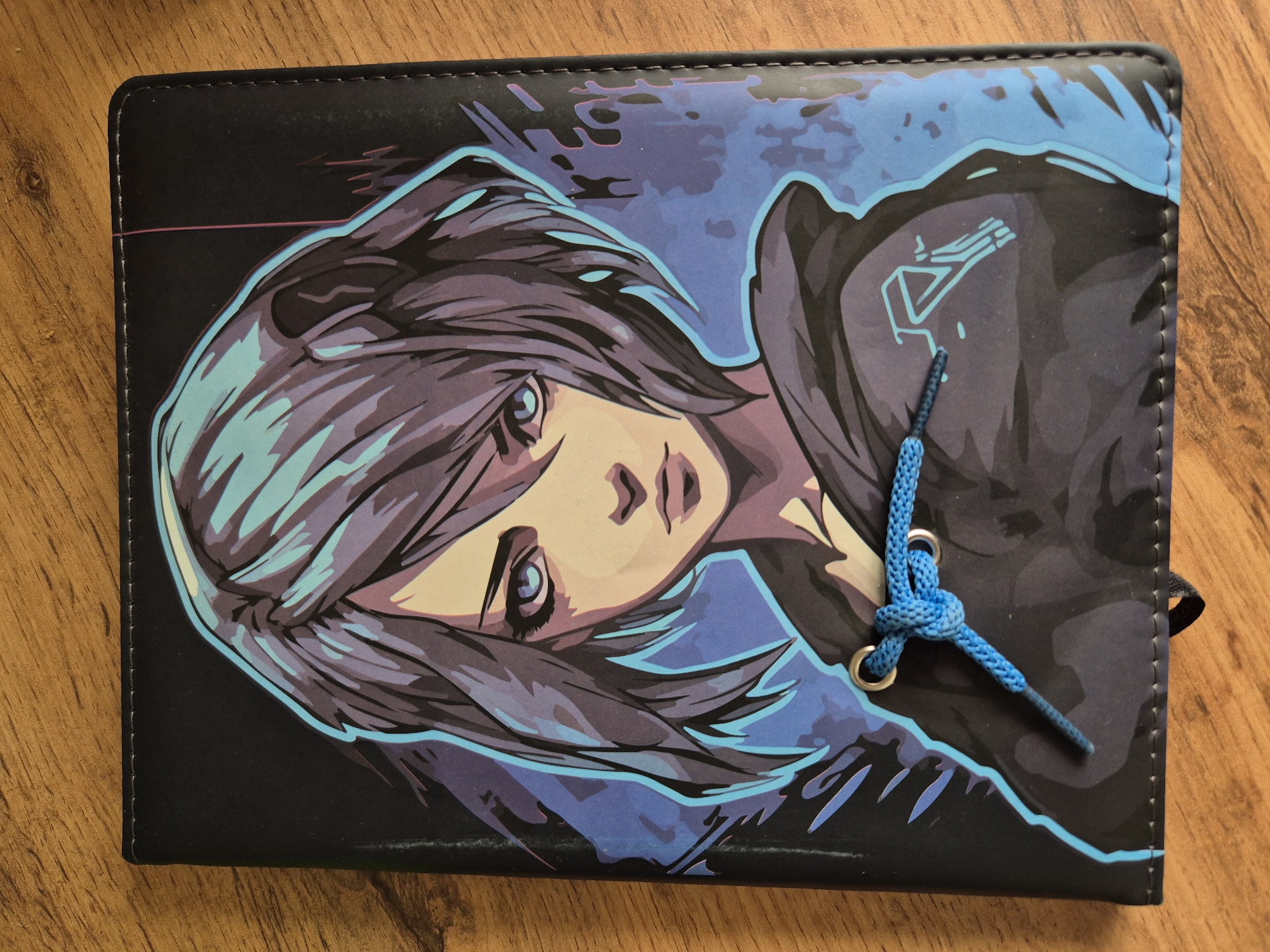 The image shows a closed notebook resting on a wooden surface. The notebook cover has a graphic design featuring an anime-style female character with short, layered hair in shades of blue and gray. She has a light complexion and is looking slightly to the left of the frame. The character is wearing a dark hooded garment, adorned with a small, stylized geometric symbol in a blue hue. The background of the graphic design is a mix of black and blue shades. A blue cord is threaded through metal eyelets on the right side of the notebook, tied in a knot. There is a black elastic band visible at the right edge of the notebook. The setting is undefined. No people are visible. The location is Unknown, Unknown.