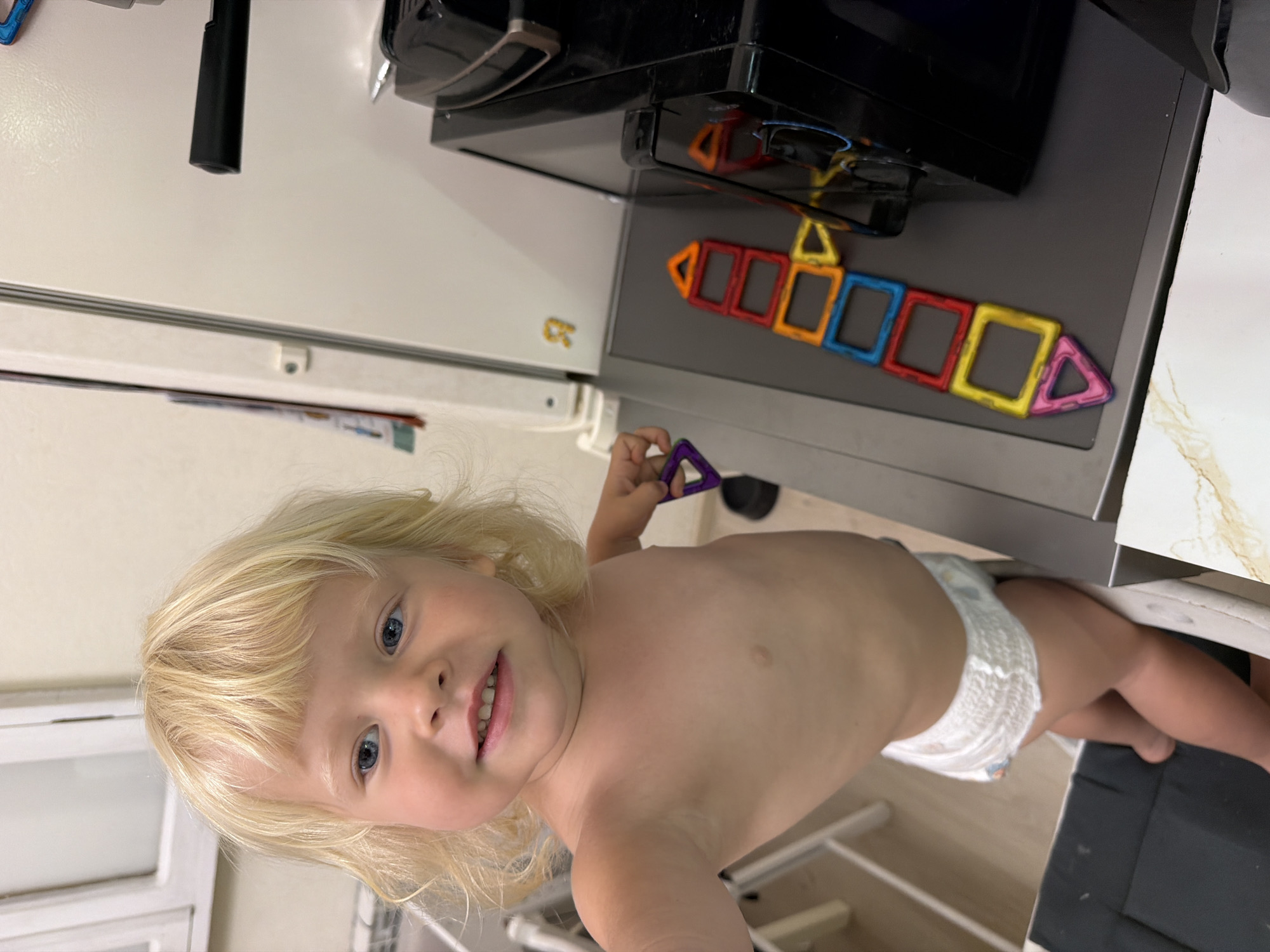 A young child with blonde hair and blue eyes is pictured from the waist up, looking towards the camera with a slight smile. The child is wearing a diaper. In the background, a magnetic toy construction set is arranged on a dark surface, resembling a colorful chain. The scene appears to be indoors, with neutral-colored walls and some shelving visible. The lighting suggests it could be daytime. The overall mood is casual and domestic.