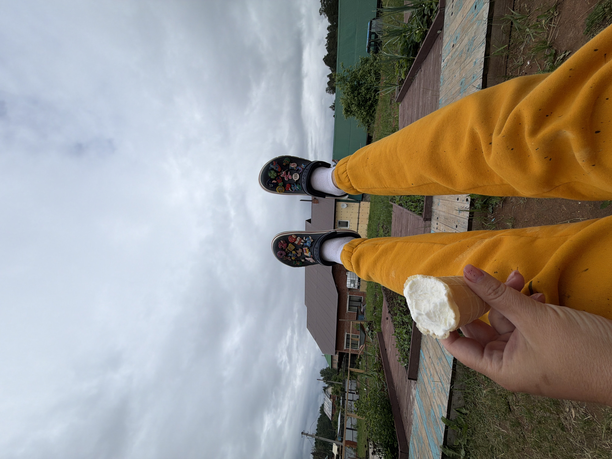 The image depicts a low-angle, first-person view of a person's lower extremities and left hand holding an ice cream cone, situated in a semi-rural environment identified as Chaikovsky, Russia. The person is wearing mustard-yellow sweatpants, white socks, and dark clogs decorated with multiple colorful charms. The left hand, exhibiting short nails, holds a waffle cone containing white ice cream, which shows signs of partial consumption.
The mid-ground features several raised garden beds constructed from weathered wood, some displaying faded blue paint, containing small green plants. The background is composed of a cloudy, overcast sky and a series of buildings: a light-colored house with a red-brown roof on the left, a brick house with a dark brown roof in the center-right, and a long building with a green corrugated metal roof further to the right. A wooden utility pole with power lines is visible on the left side of the background, and a blue vehicle is partially visible behind the green-roofed building. The immediate foreground and areas around the garden beds consist of sparse grass and soil.
