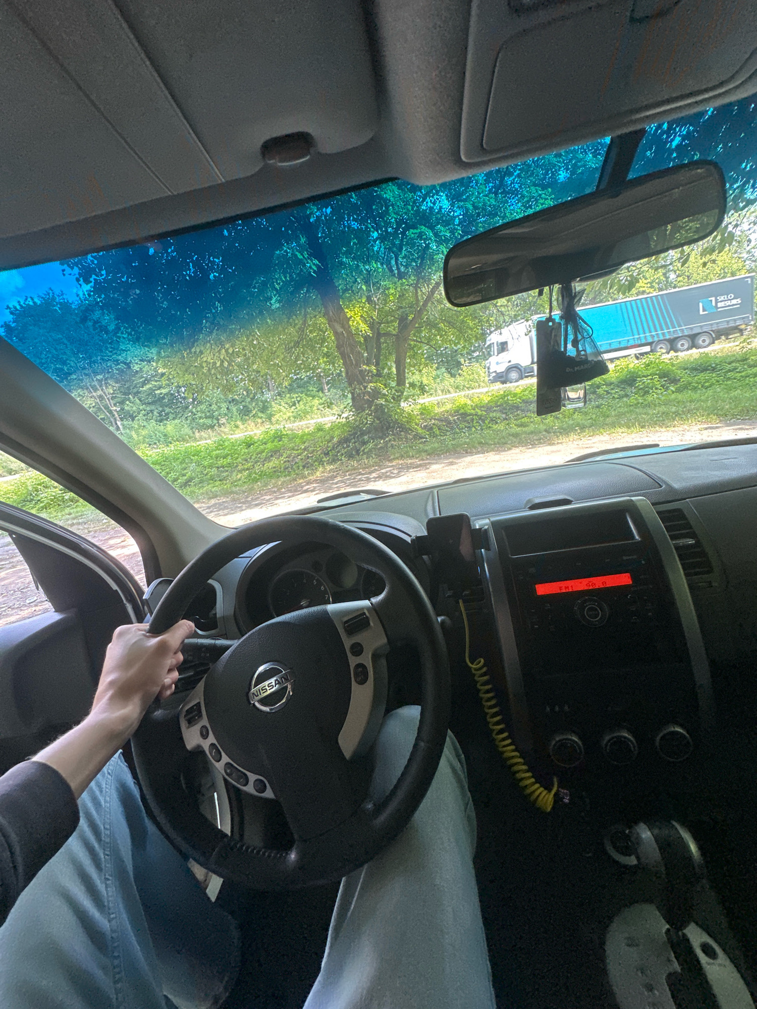 The image is taken from the driver's perspective inside a car, looking forward and slightly to the right. The car's interior is visible, including the steering wheel with a Nissan logo, the dashboard with a car radio displaying "FM 98.8", and a gear shifter. A person's hands are on the steering wheel, and their legs are clad in light blue denim. Outside the car, through the windshield, lush green trees and vegetation are visible, suggesting a rural or semi-rural environment. A large white truck with a blue trailer is seen driving on a road in the distance. The setting appears to be the city of Shatava, Ukraine, based on the provided context. The lighting suggests it is daytime.