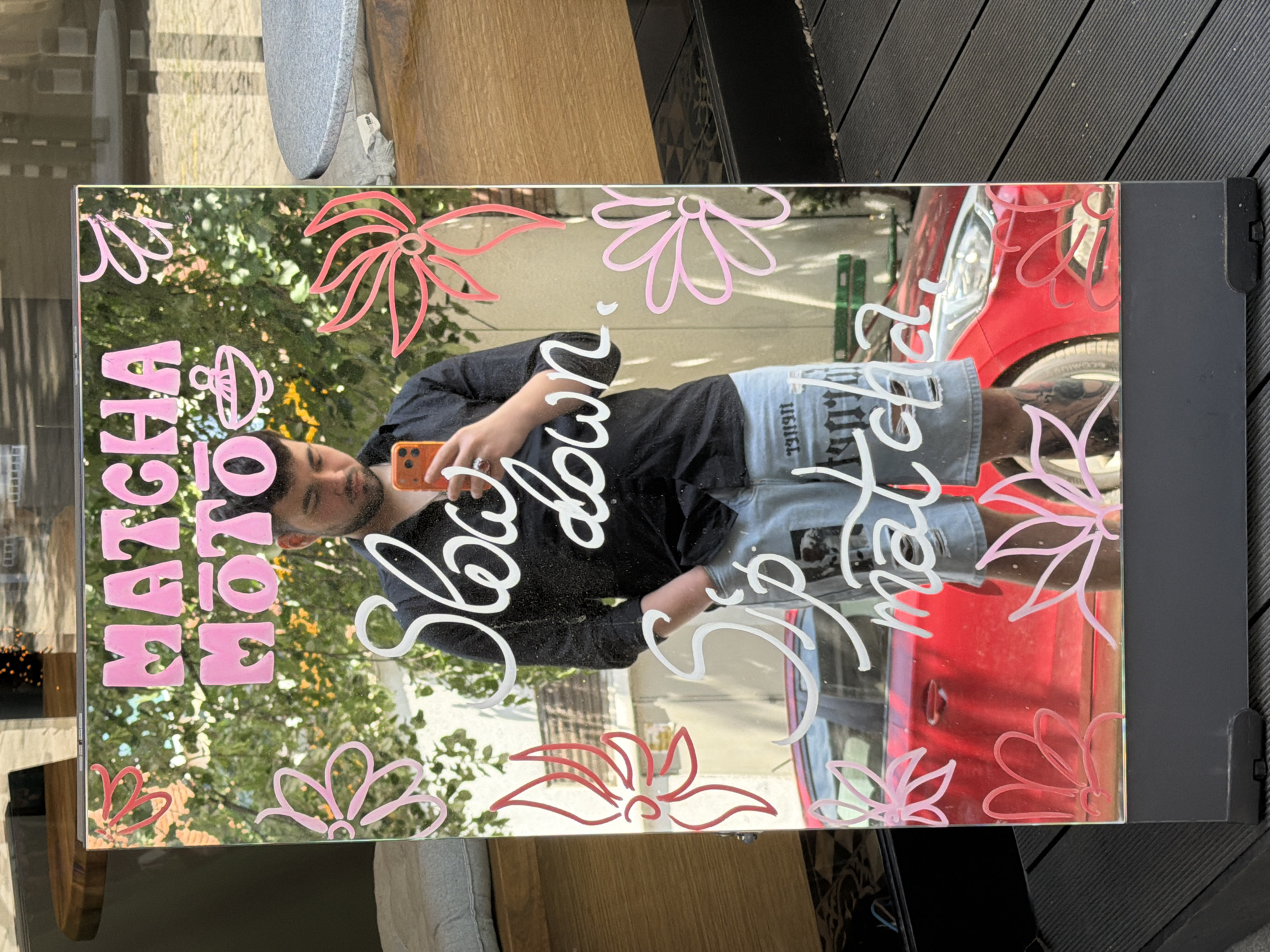 The image shows a rectangular mirror with decorative elements, reflecting a man taking a selfie with a mobile phone. The mirror has pink lettering that reads "MATCHA MOTO" and white script that reads "Slow down Sip matcha". Decorative pink and red flower drawings are also present on the mirror's surface. The man is wearing a black top and light denim shorts. The reflection also shows a red car and foliage, suggesting an outdoor setting. The mirror is resting on an outdoor table, with a wooden surface and a black composite material with a slatted texture in the background. There is insufficient detail to identify a specific location in Sofia, Bulgaria.